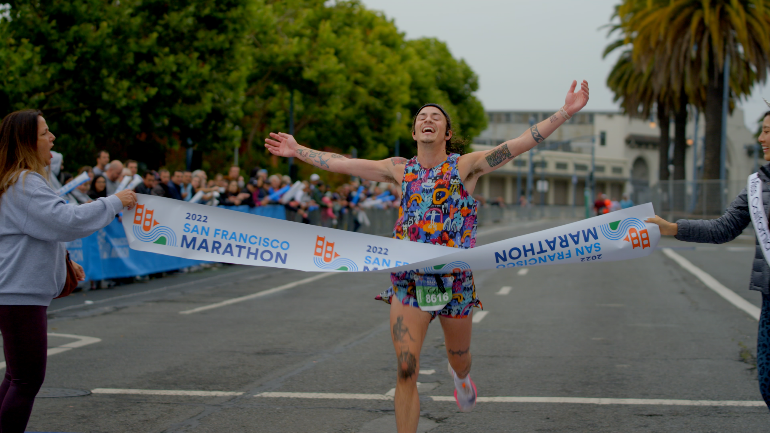 A runner joyfully crosses the finish line of the 2022 San Francisco Marathon, arms raised in triumph, cheered by onlookers holding the finishing tape.