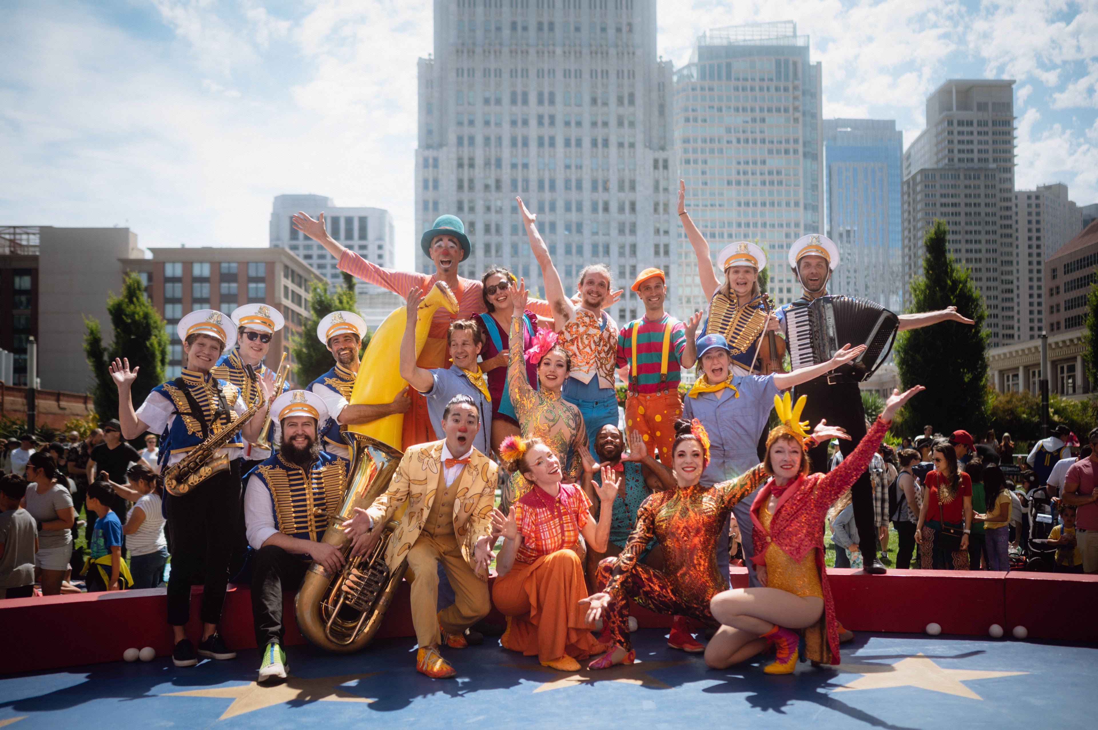 A lively group of performers in colorful costumes and hats pose energetically in front of a cityscape, with some playing musical instruments.