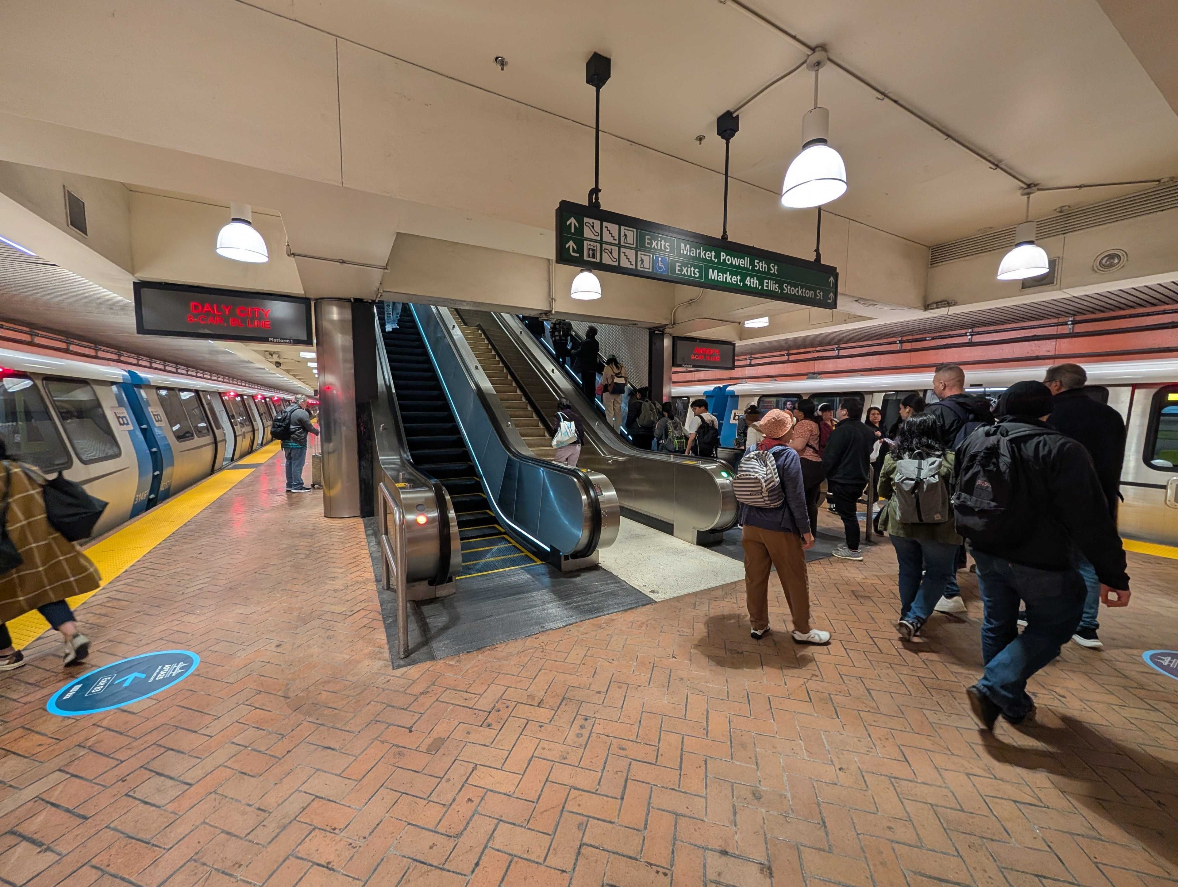 People crowd on a subway platform with trains on both sides. An escalator leads up, and signs indicate exits to Market, Powell, and other streets.
