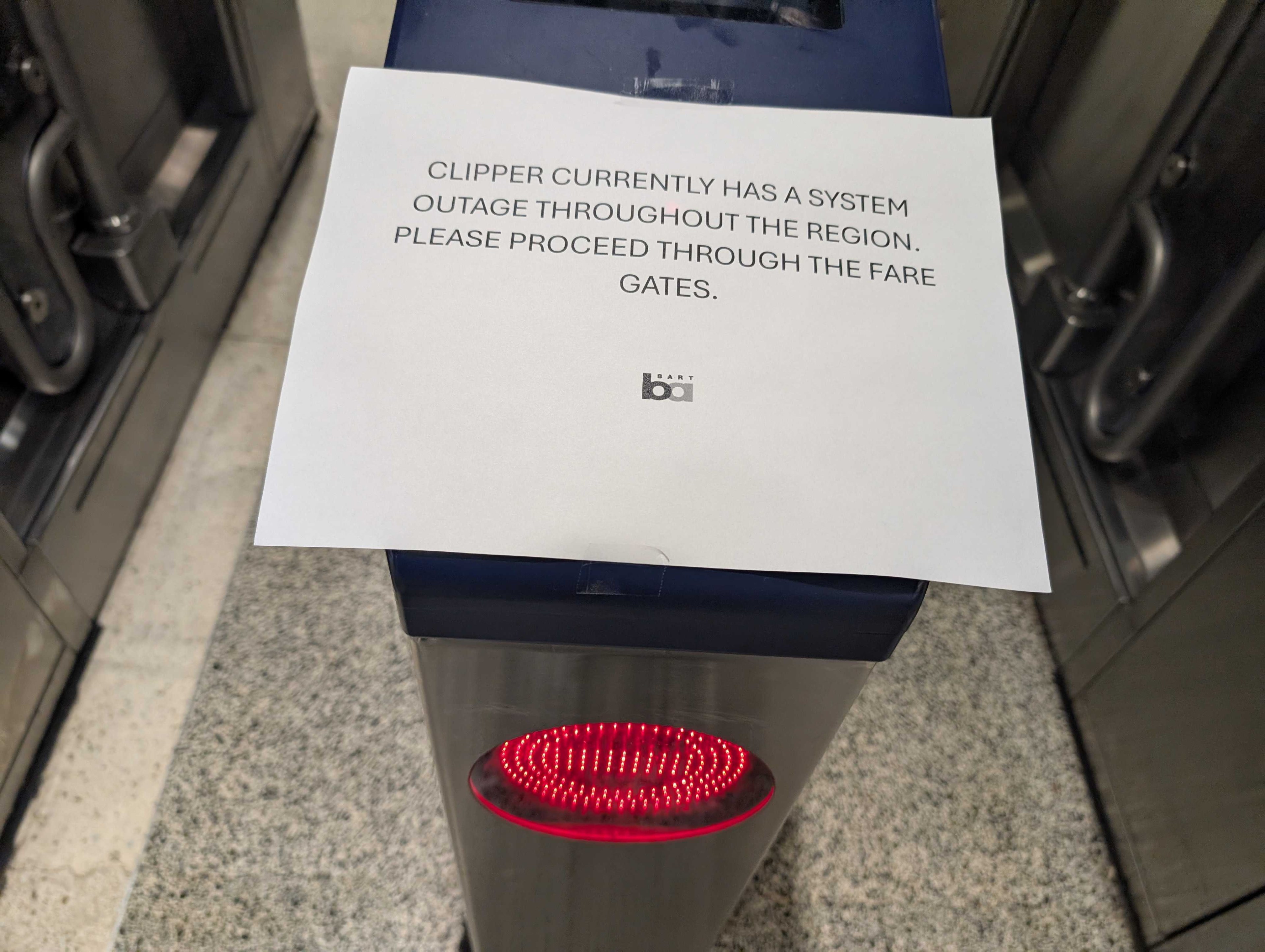A sign on a transit fare gate reads, &quot;Clipper currently has a system outage throughout the region. Please proceed through the fare gates.&quot;