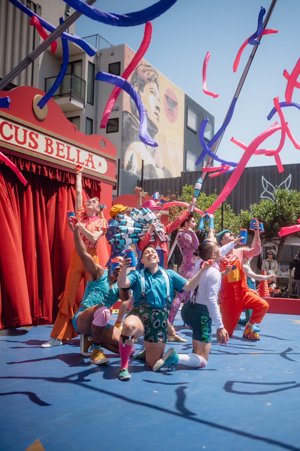 A group of performers in colorful costumes joyfully pose in front of a red circus tent with streamers flying above them, under a clear blue sky.