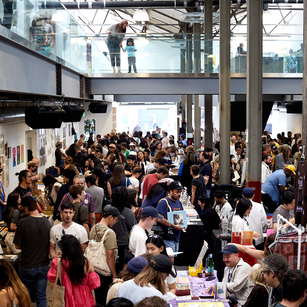 A busy indoor market or fair with crowded tables displaying various items. People are interacting, and a glass upper level shows a few more observers.