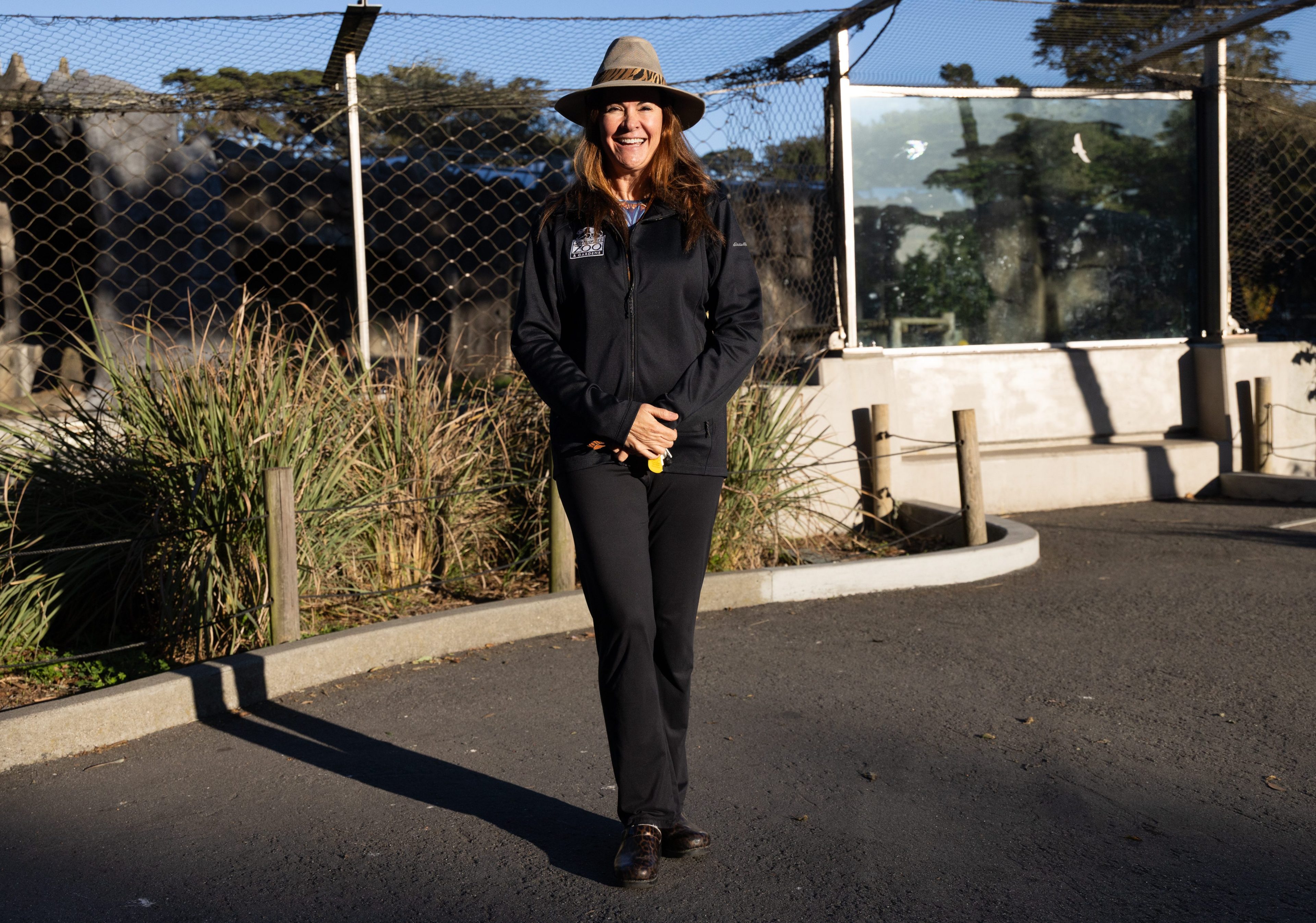 A smiling person stands on a paved path, wearing a wide-brimmed hat and dark clothing. They are in front of a fenced area with plants and trees.