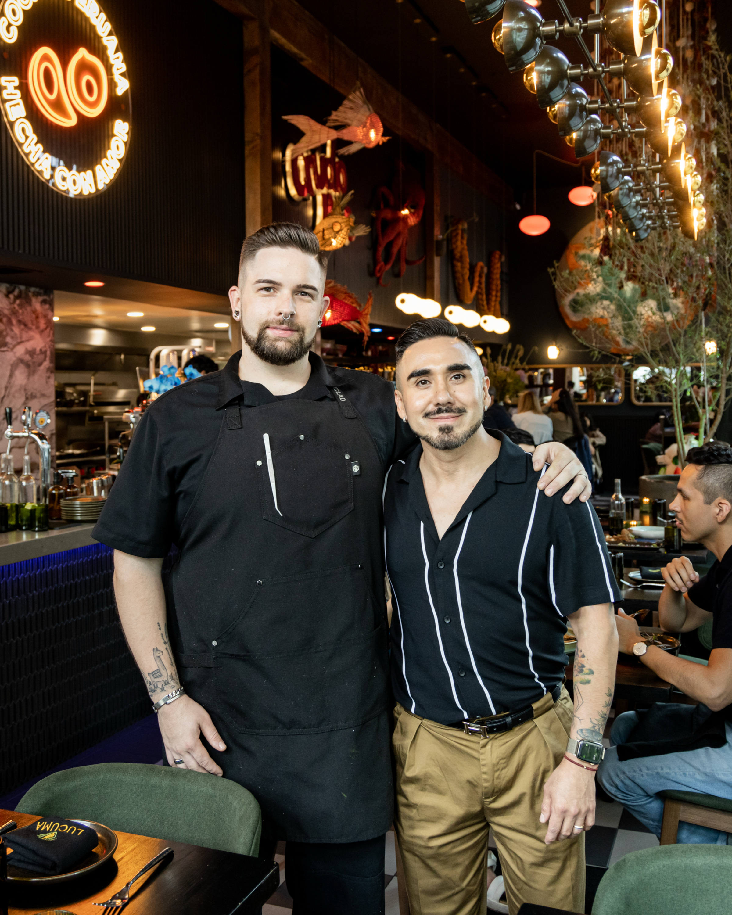 Two people stand smiling in a vibrant restaurant. One wears a black apron, the other a black striped shirt with tan pants. Decorative lights and plants fill the space.