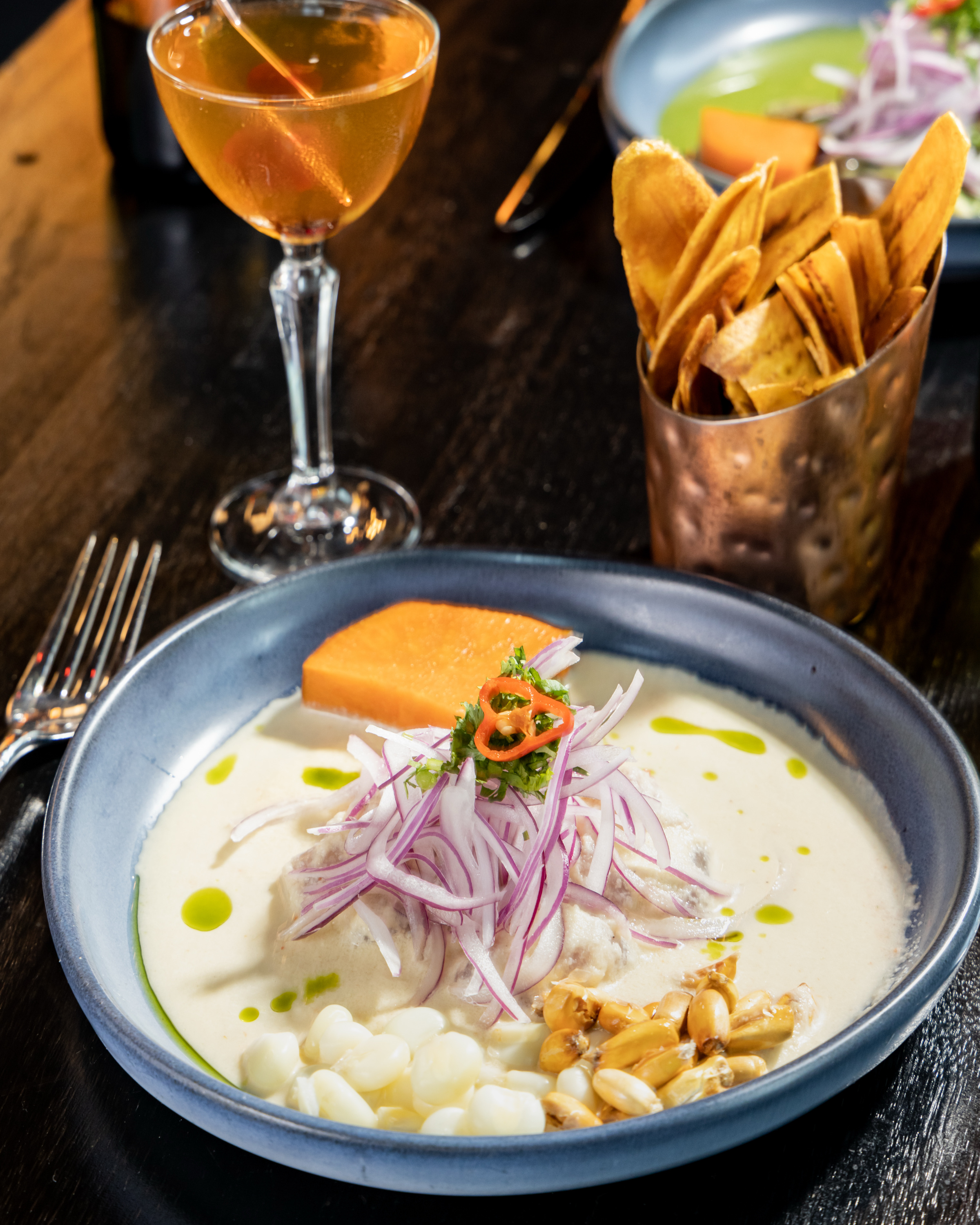 The image shows a plate of ceviche with white sauce, sliced red onions, corn, and sweet potato. A crispy snack is in a cup, and there's a cocktail nearby.