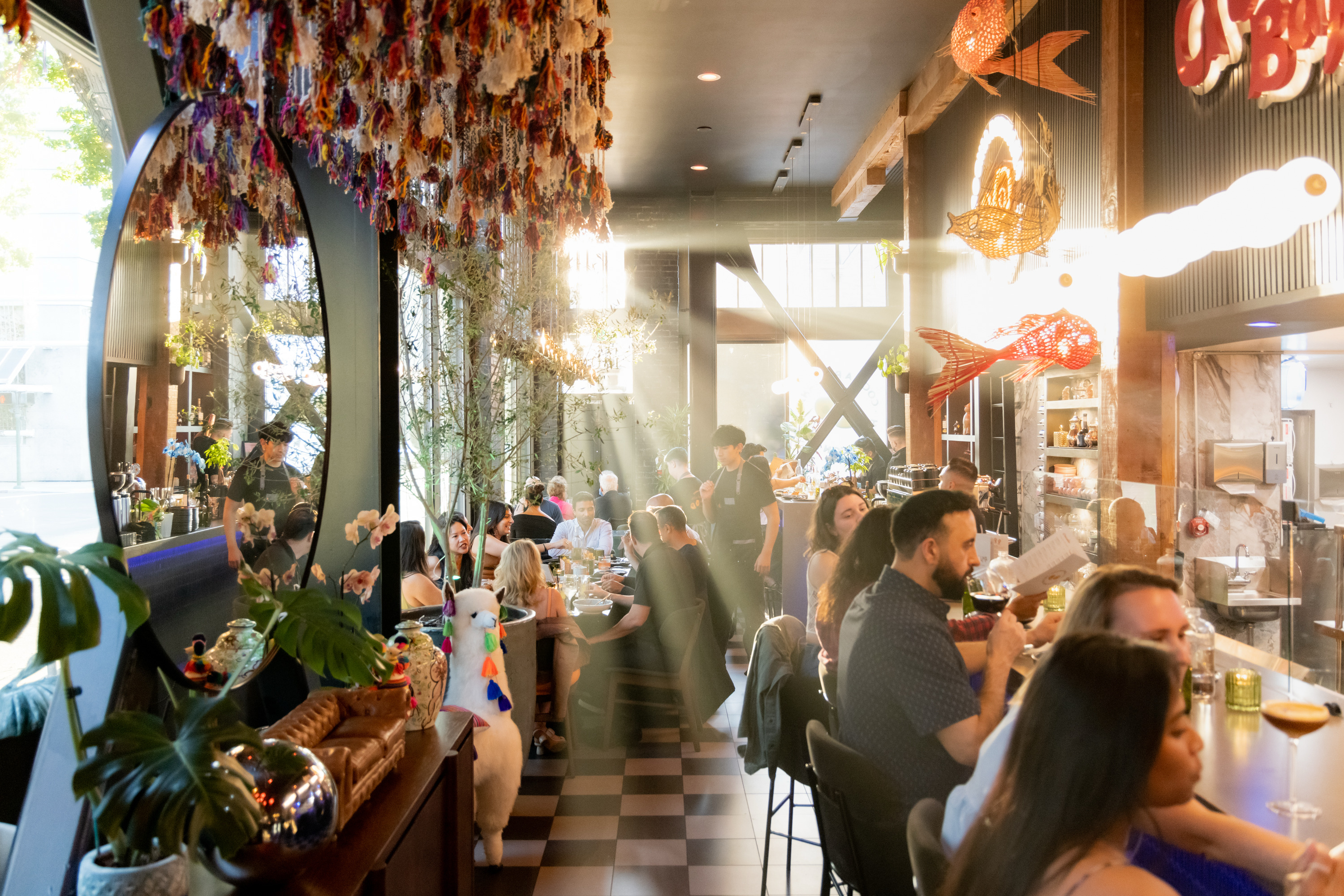 The image shows a bustling restaurant with people dining. Sunlight streams through large windows, illuminating hanging decorations and a lively atmosphere.