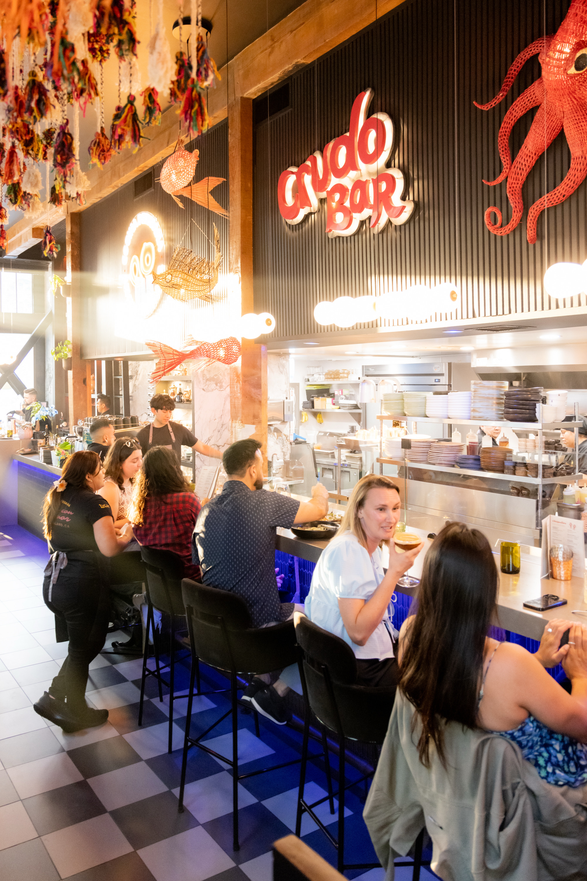 The image shows a lively bar with people seated on stools at a counter. The decor includes colorful fish and an octopus, with a sign saying "Crudo Bar".