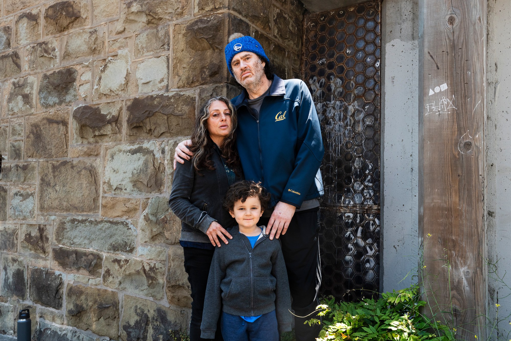 A family of three stands against a stone wall. The adults look serious while the child smiles. The man wears a blue jacket and beanie, and the woman wears a dark jacket.