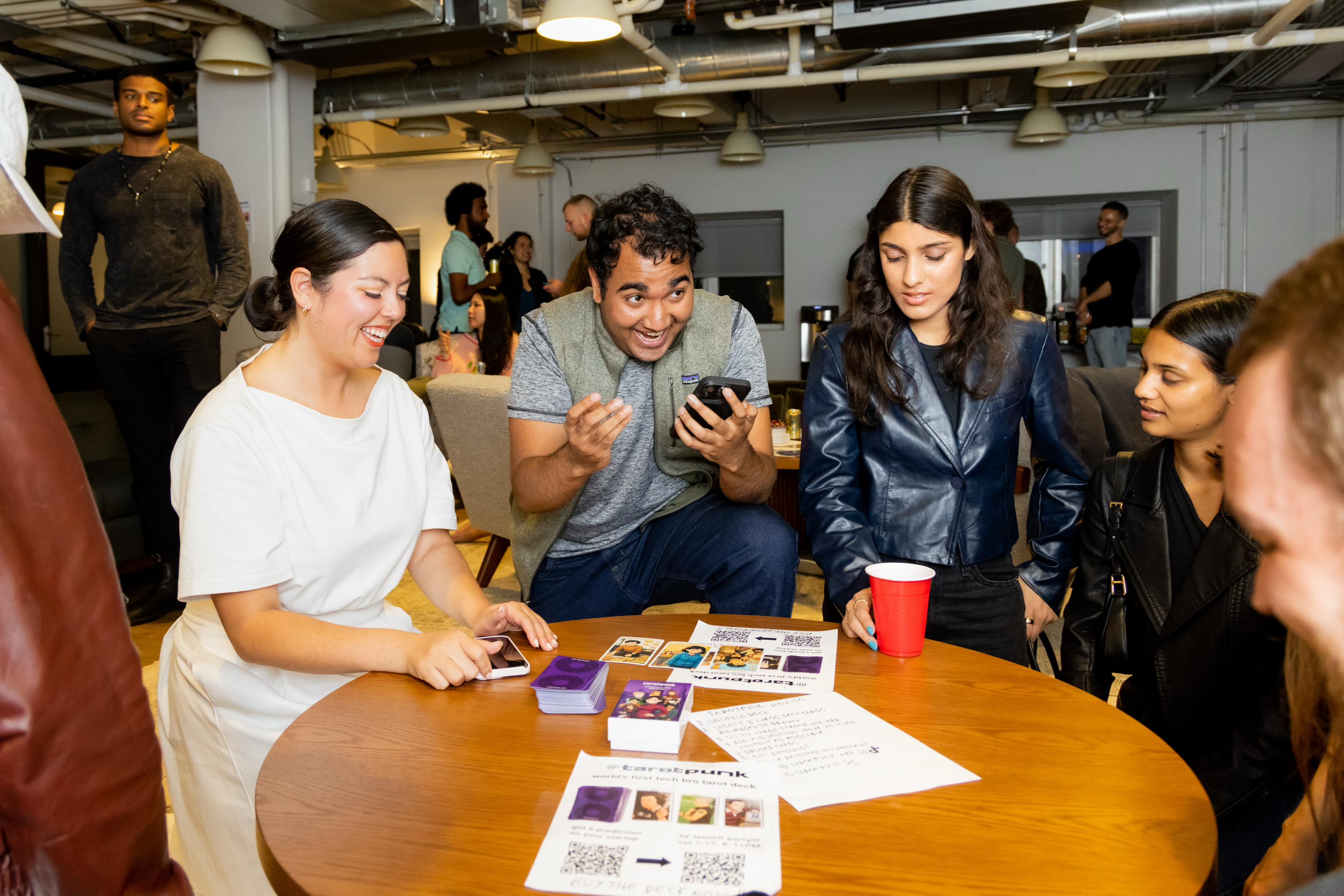 Several people gather around a table, looking at a phone. A woman smiles, and others are engaged. Papers and QR codes are on the table, and a red cup is nearby.