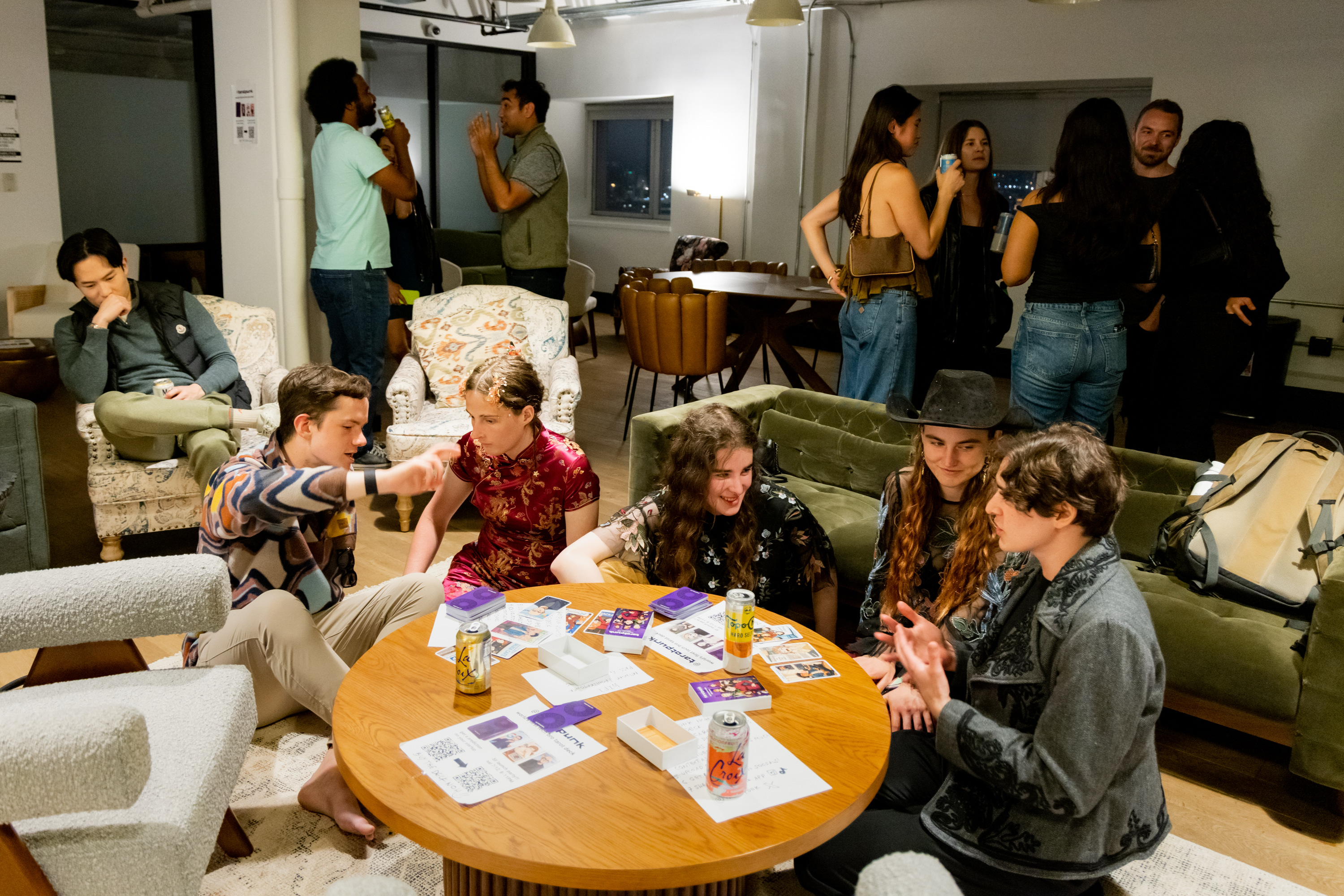 People are gathered in a cozy room. Some are seated around a table with cards and drinks, while others stand and chat in the background.