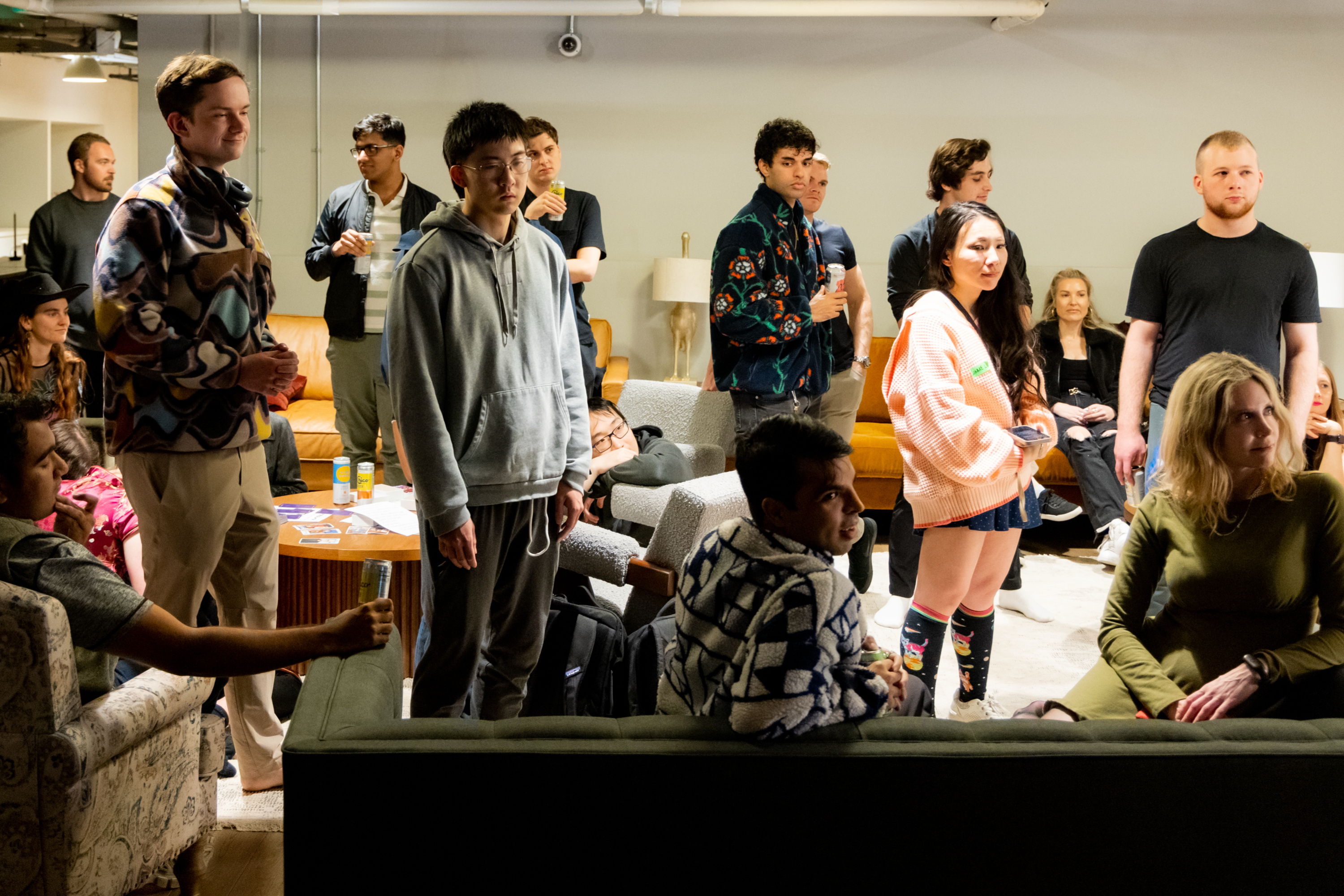 A group of people, some standing and others sitting on sofas, are gathered in a well-lit room. They're casually dressed, engaged, and focused on something ahead.