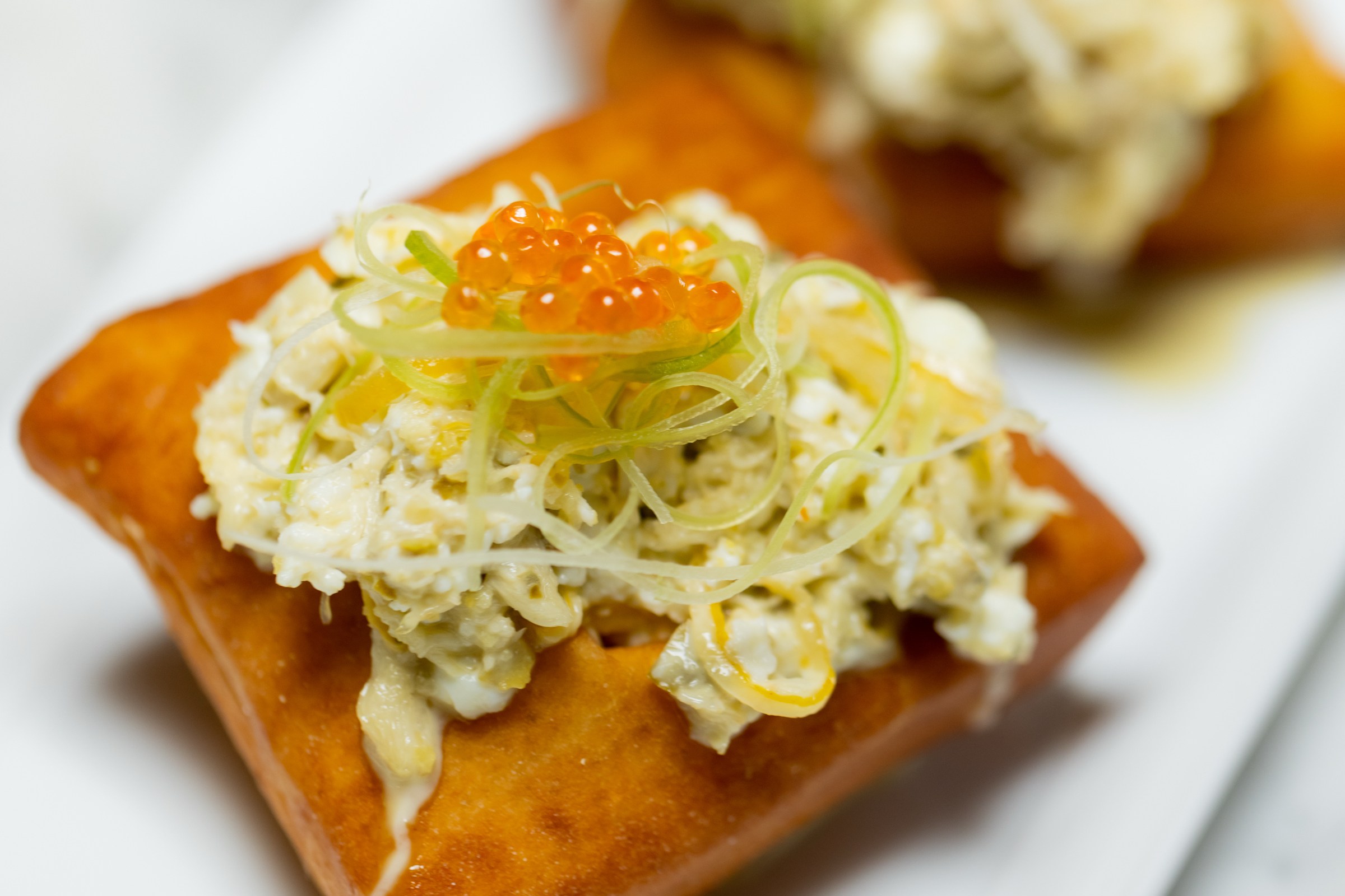 Two golden-brown pastries topped with a creamy mixture, garnished with green slivers and small orange spheres resembling fish roe, on a white plate.