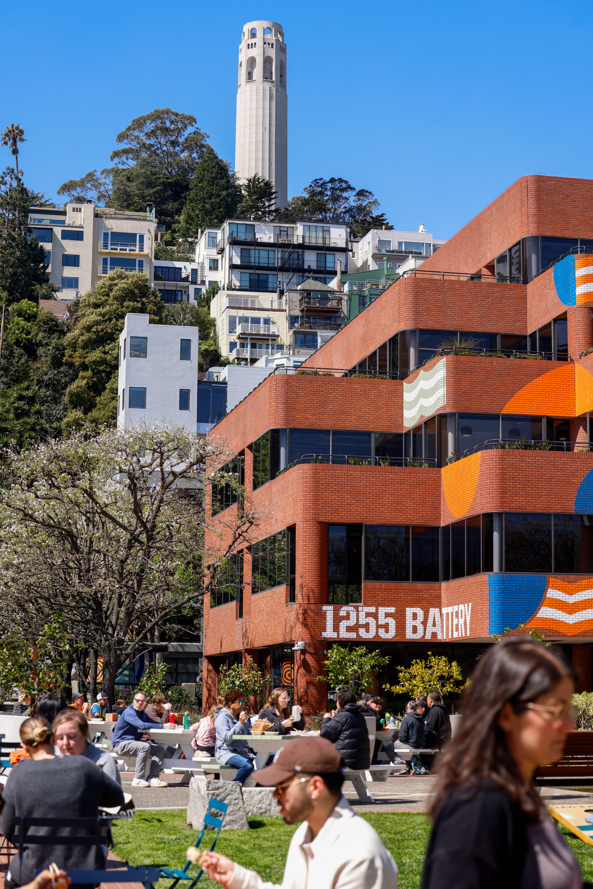 People are sitting outside a brick building with "1255 Battery" on it. Behind, a tall white tower and several houses are on a tree-covered hill under a blue sky.