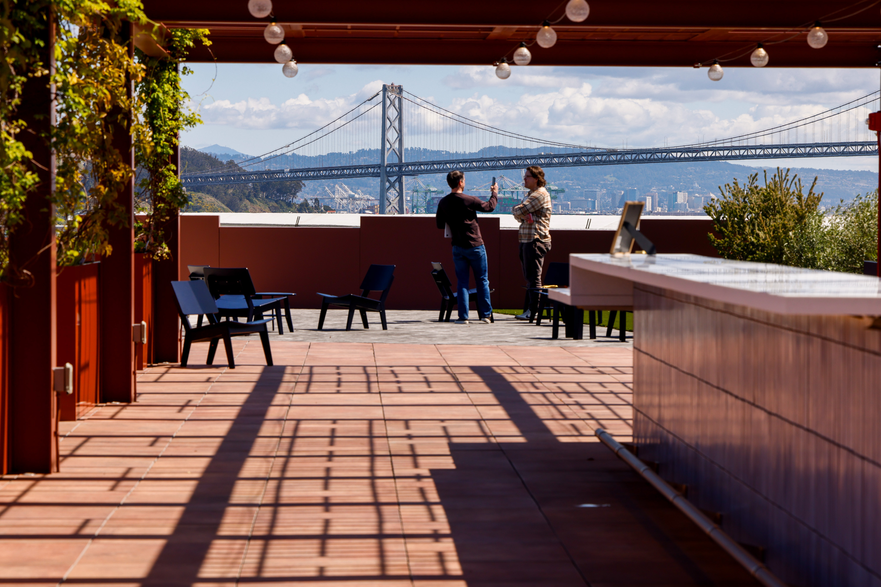 A rooftop patio with two people talking, surrounded by chairs and plants. The Bay Bridge is visible in the background under a partly cloudy sky.