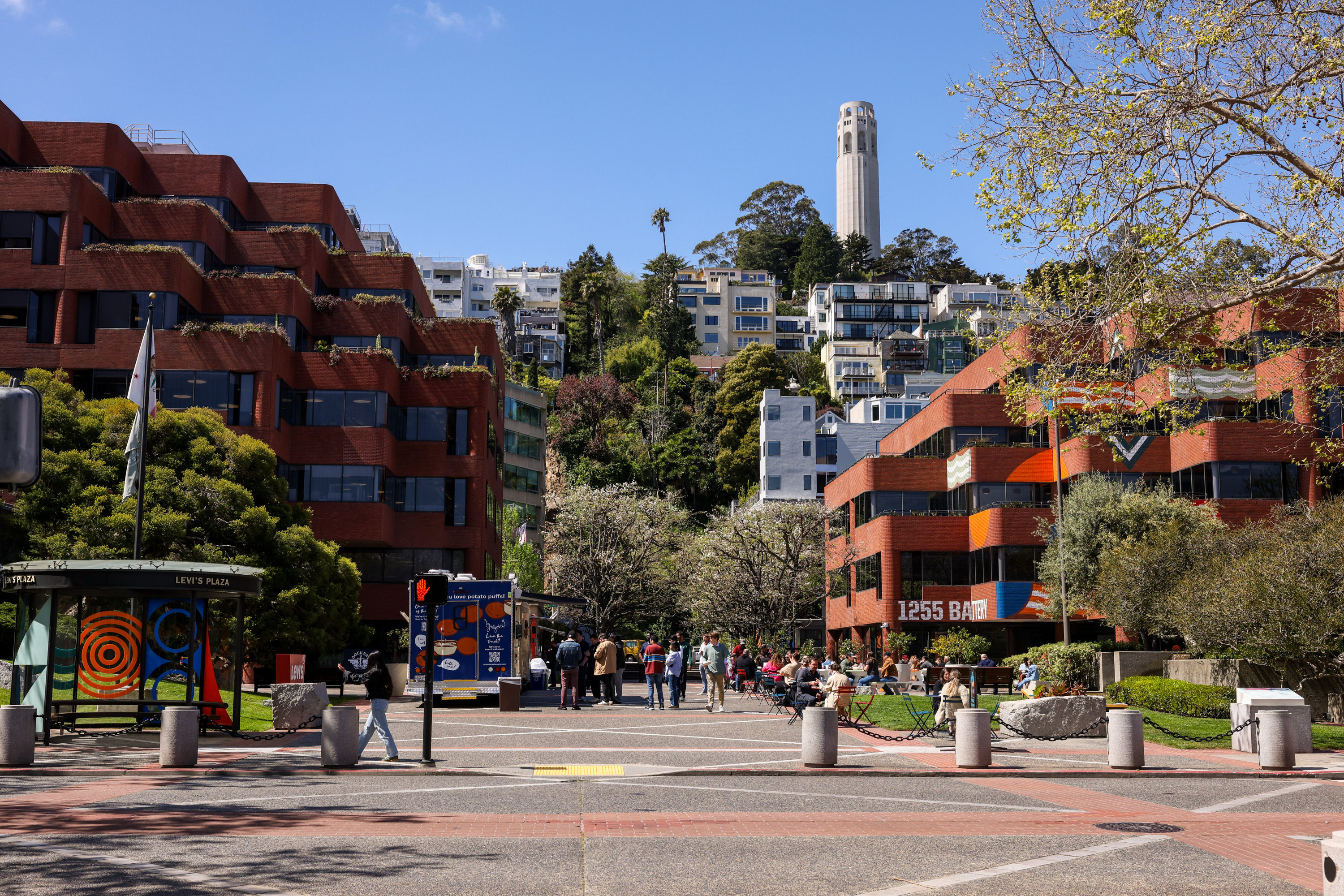 The image shows a bustling plaza with brick buildings and people relaxing. In the background, there's a hill with houses and a tall tower against a blue sky.