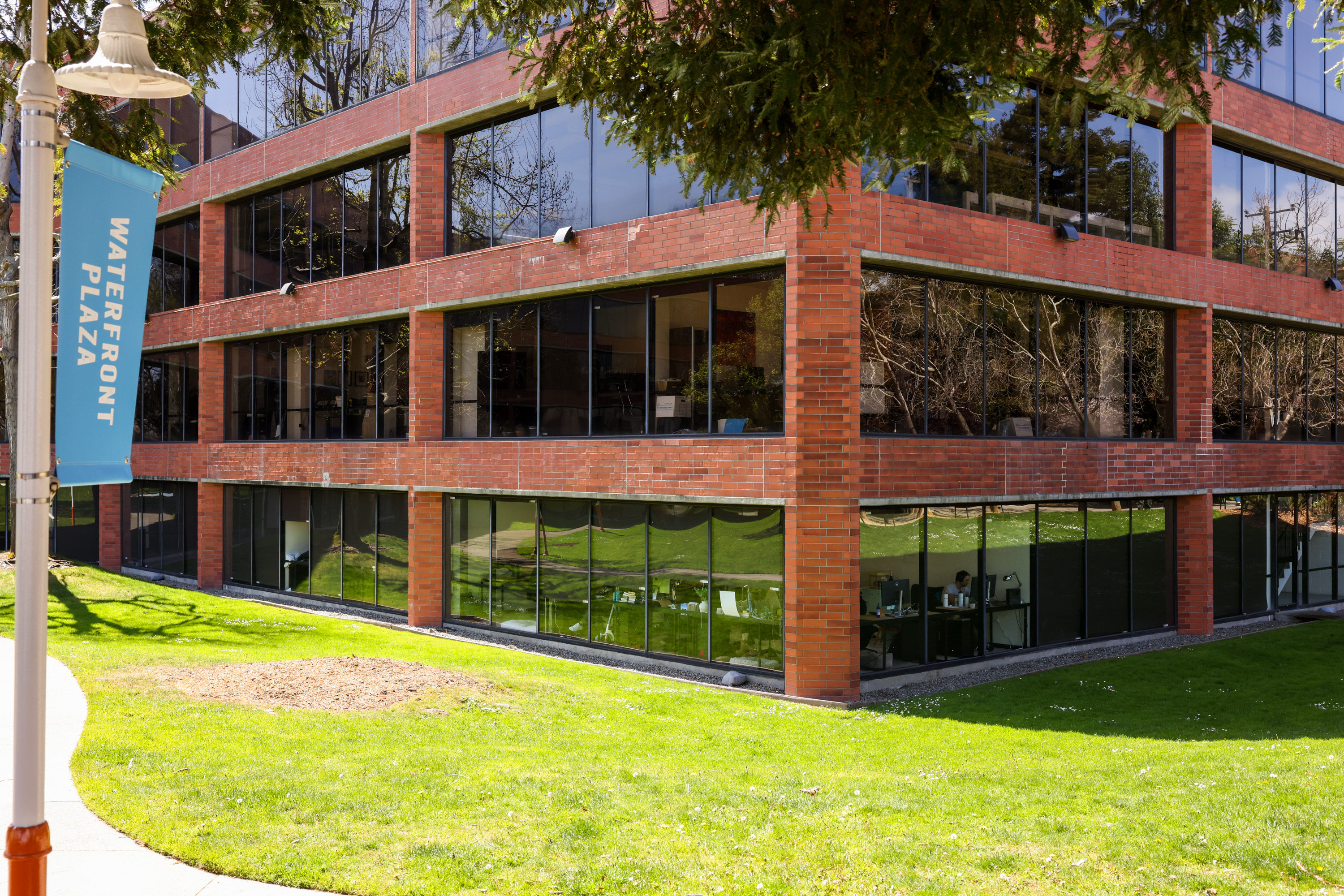 A red-brick office building with large windows is surrounded by green grass. A light blue &quot;Waterfront Plaza&quot; banner hangs on a pole in the foreground.