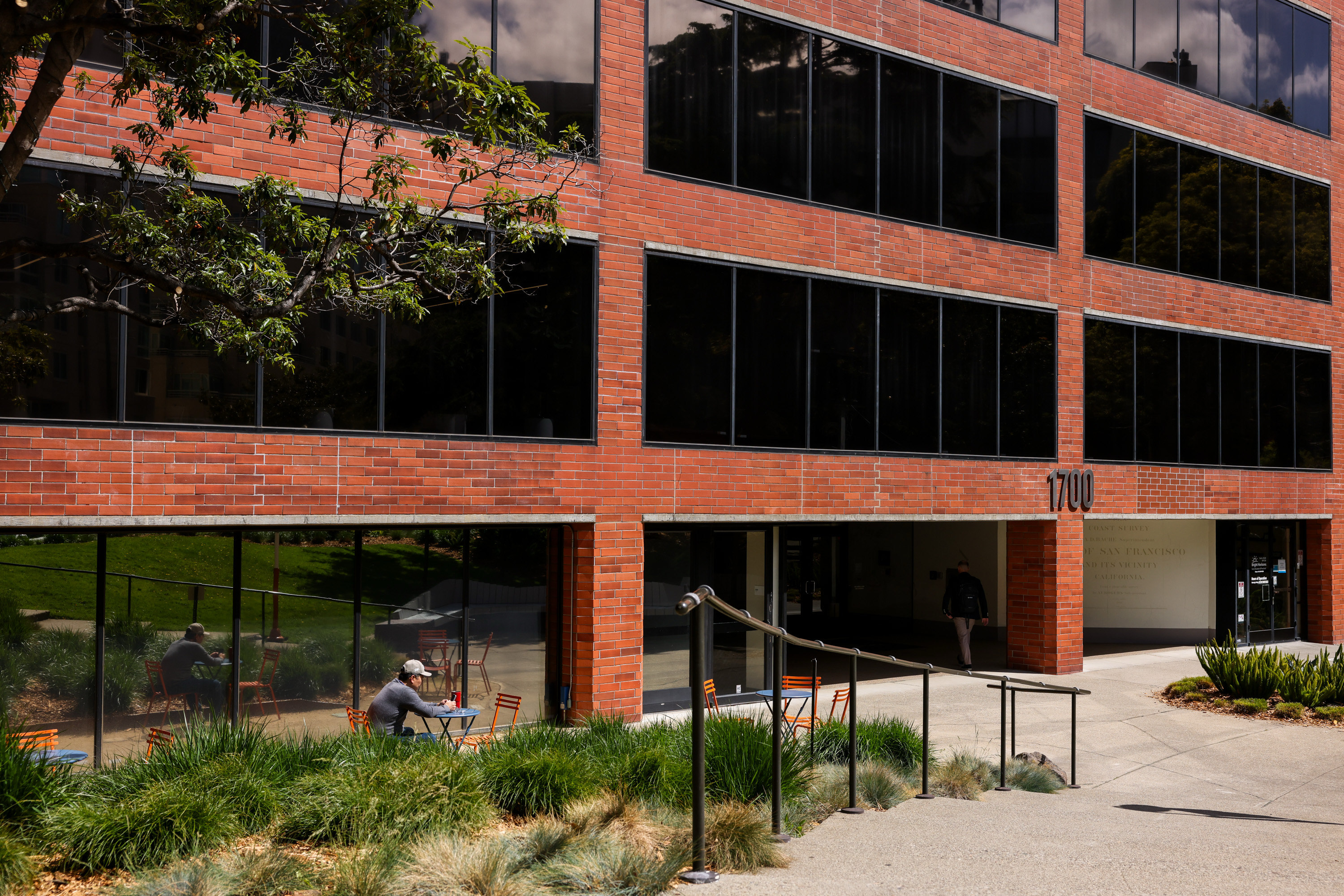 A modern brick building with dark windows stands behind a garden. A person sits at an outdoor table, and another walks into a shaded entrance labeled &quot;1700.&quot;