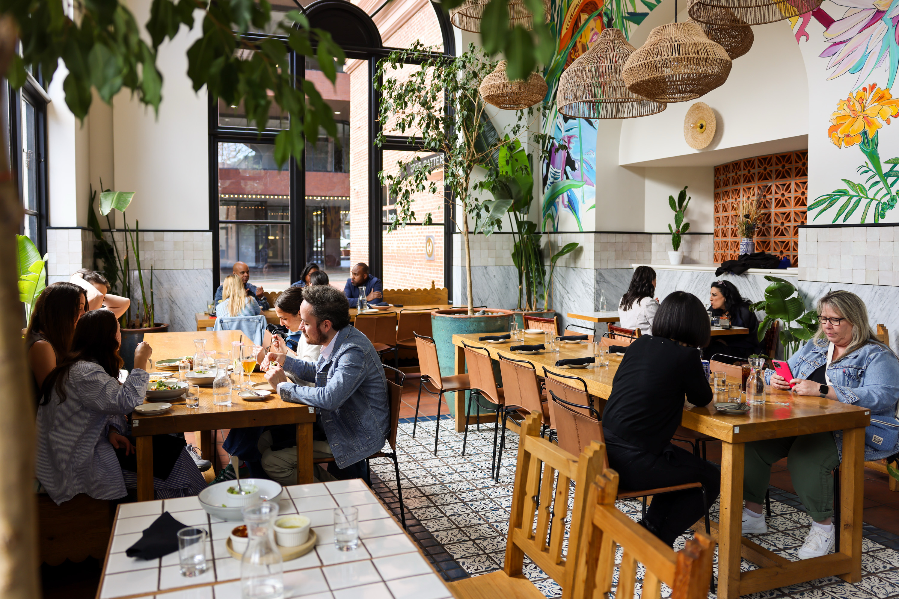 The image shows a bright, lively restaurant interior with people dining at wooden tables surrounded by plants and colorful wall art.