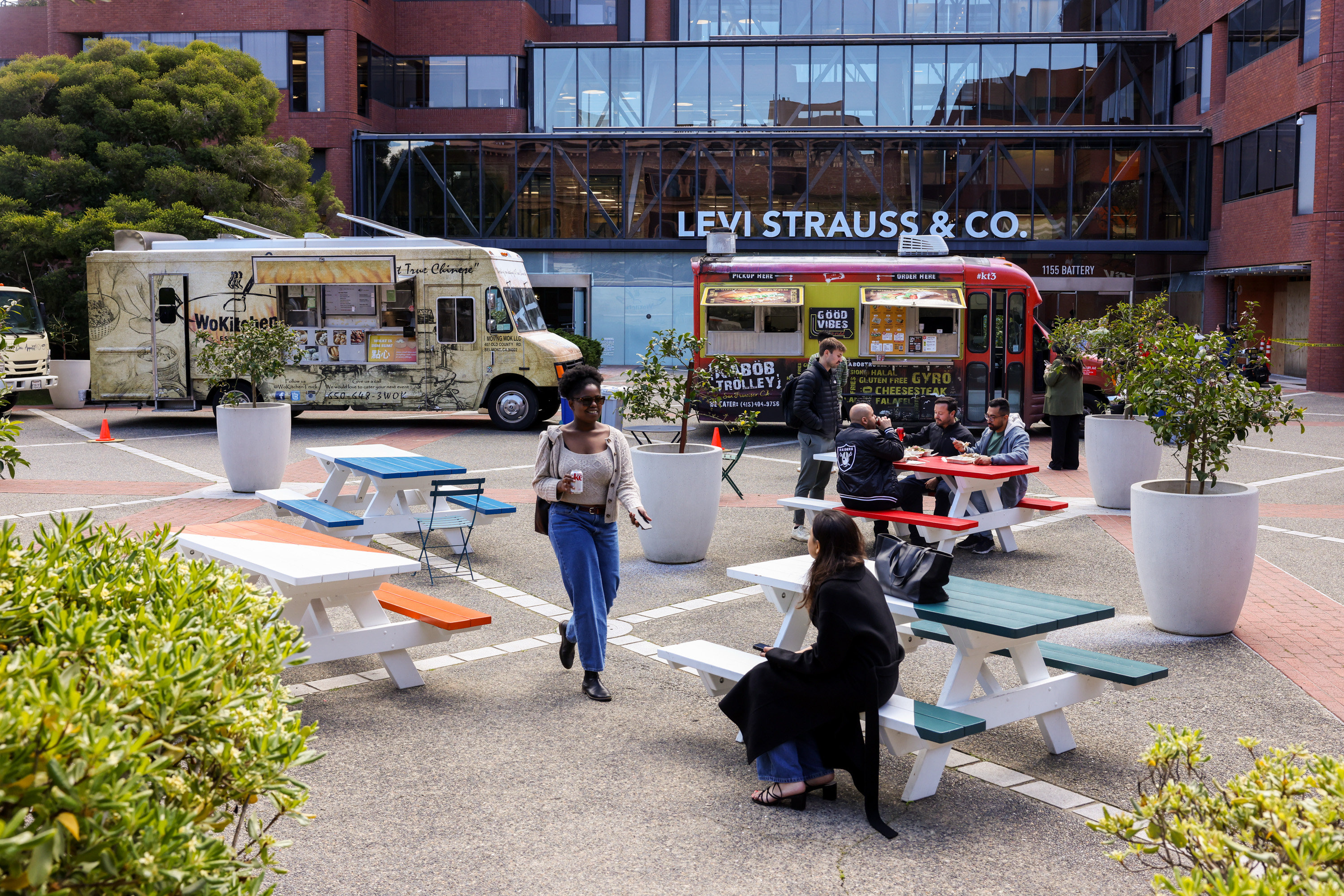A courtyard with benches has two food trucks. People sit on benches or walk, surrounded by large potted plants. The Levi Strauss &amp; Co. building is in the background.