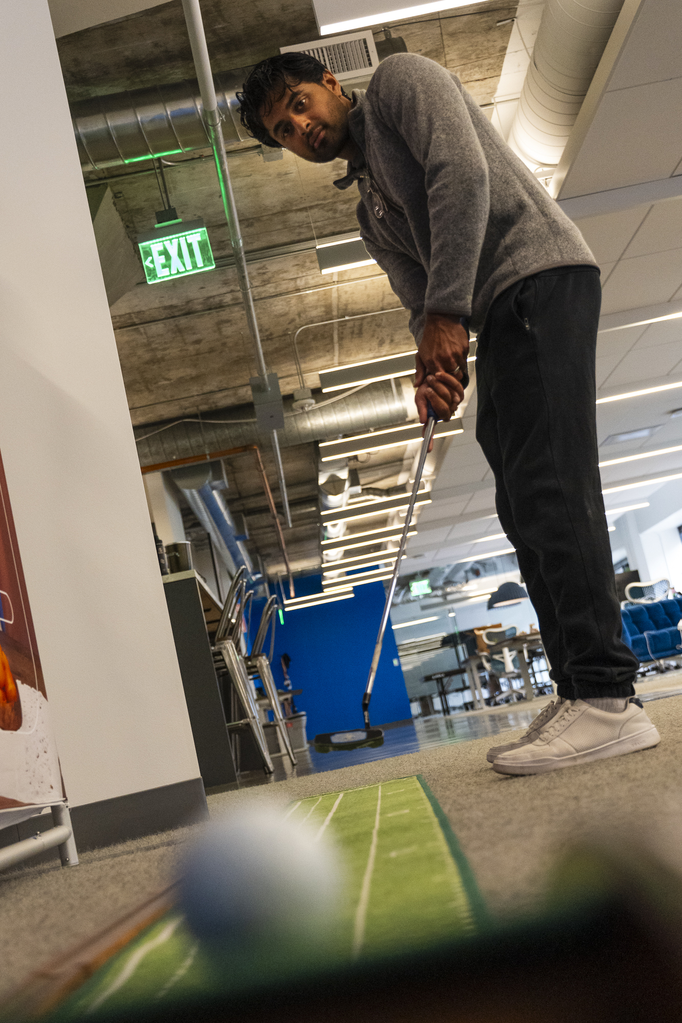 A person in a gray sweater is putting a golf ball on an indoor mat in an office with industrial ceilings, a green EXIT sign, and blue accents.