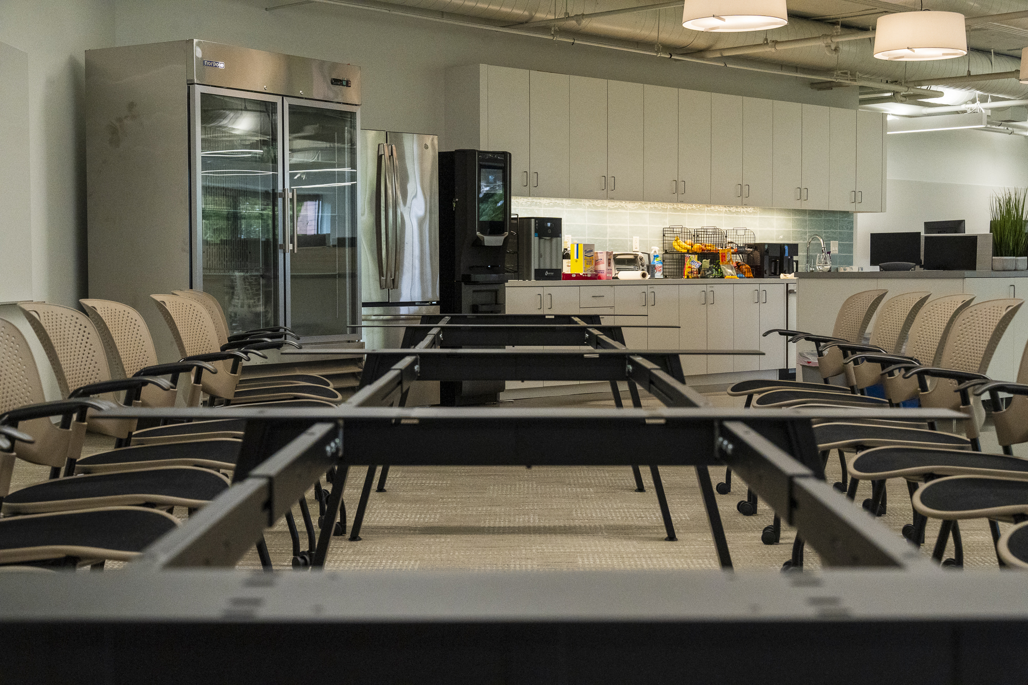 The image shows a room with chairs arranged around tables, facing a kitchenette area. The kitchenette has a fridge, coffee machine, snacks, and cabinets.