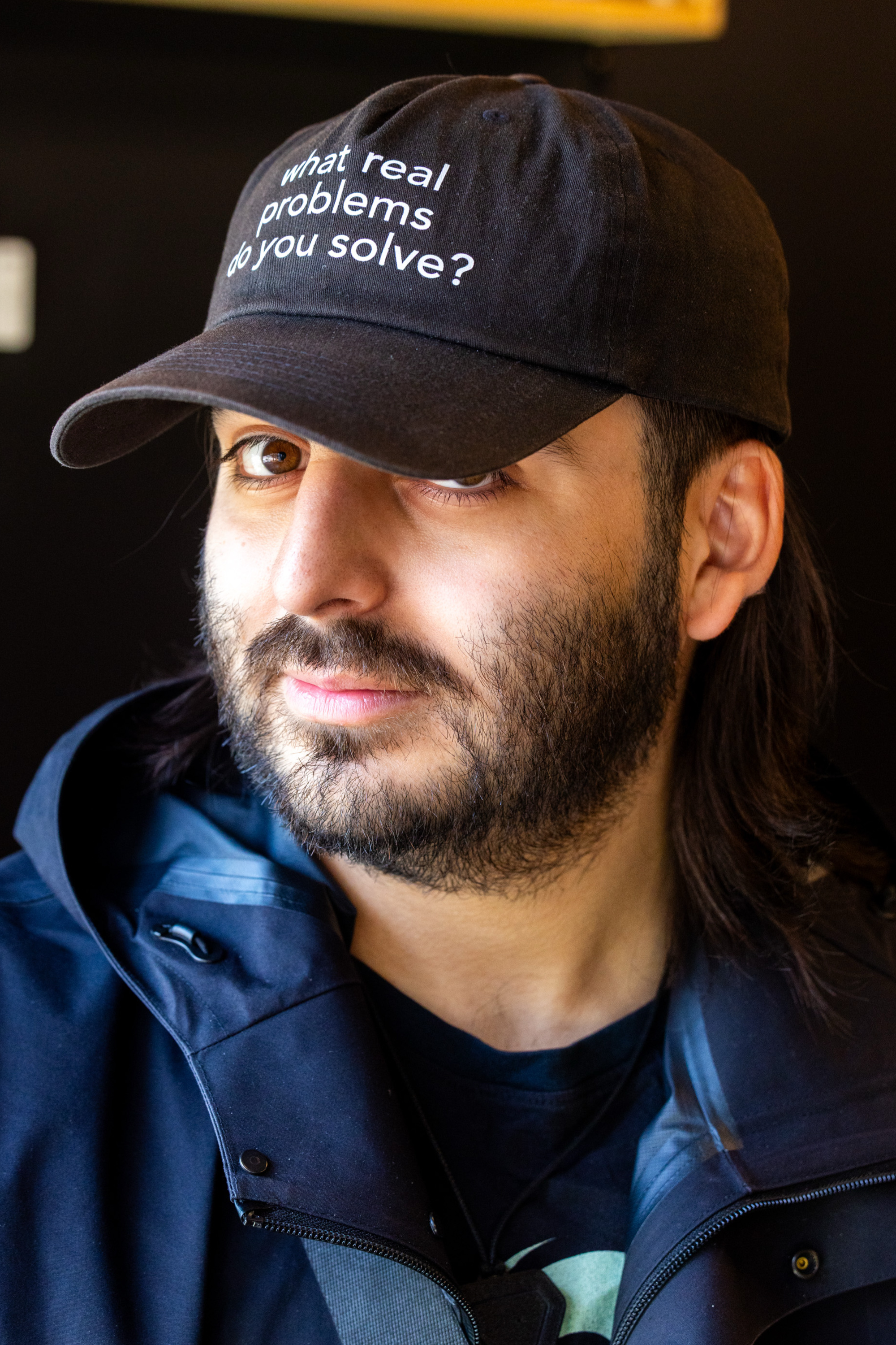 A person with long hair and a beard wears a black cap with the phrase "what real problems do you solve?" and a dark jacket, against a dark background.