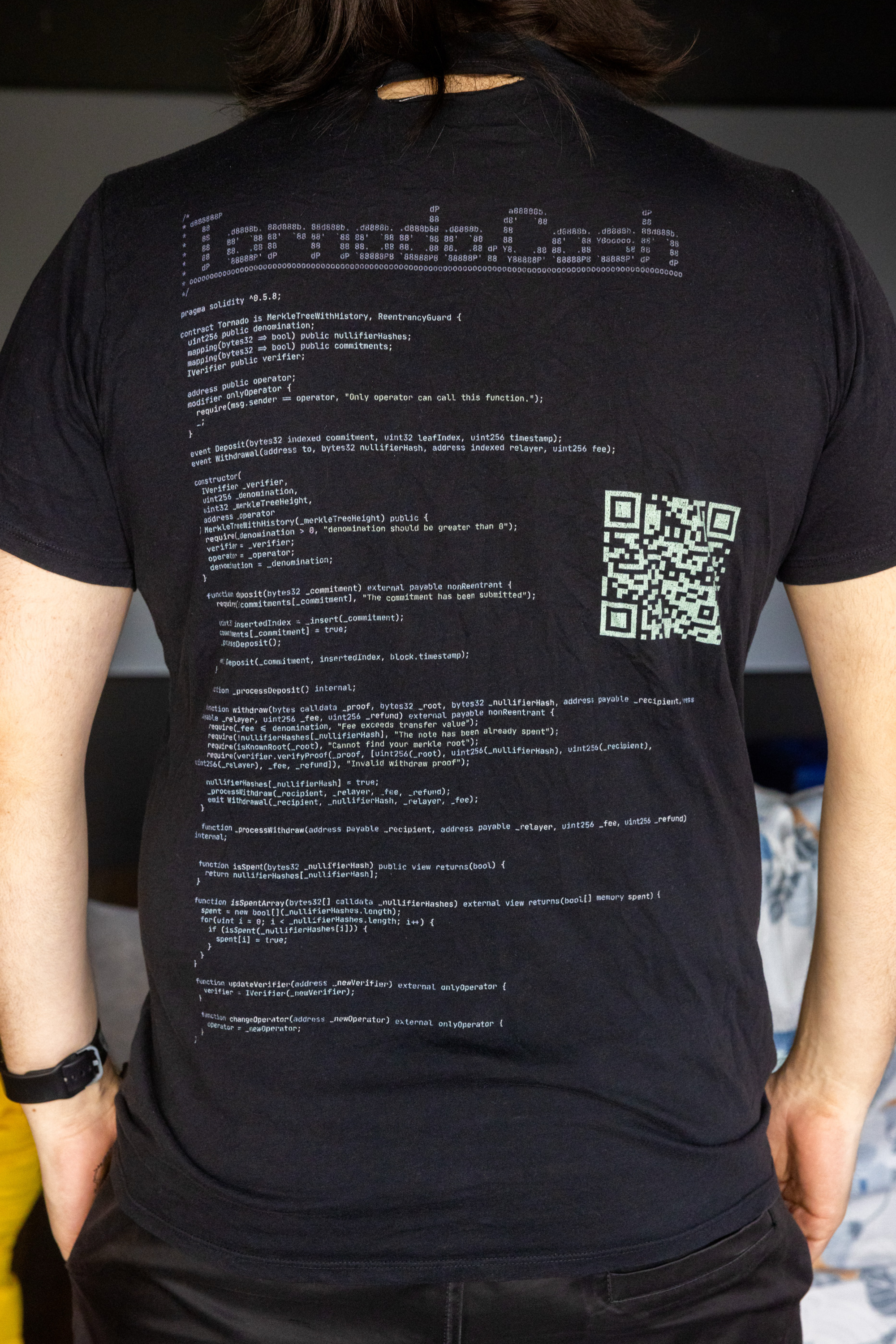 The image shows a person wearing a black t-shirt with code printed on the back. The text includes programming scripts and a QR code on the right side.