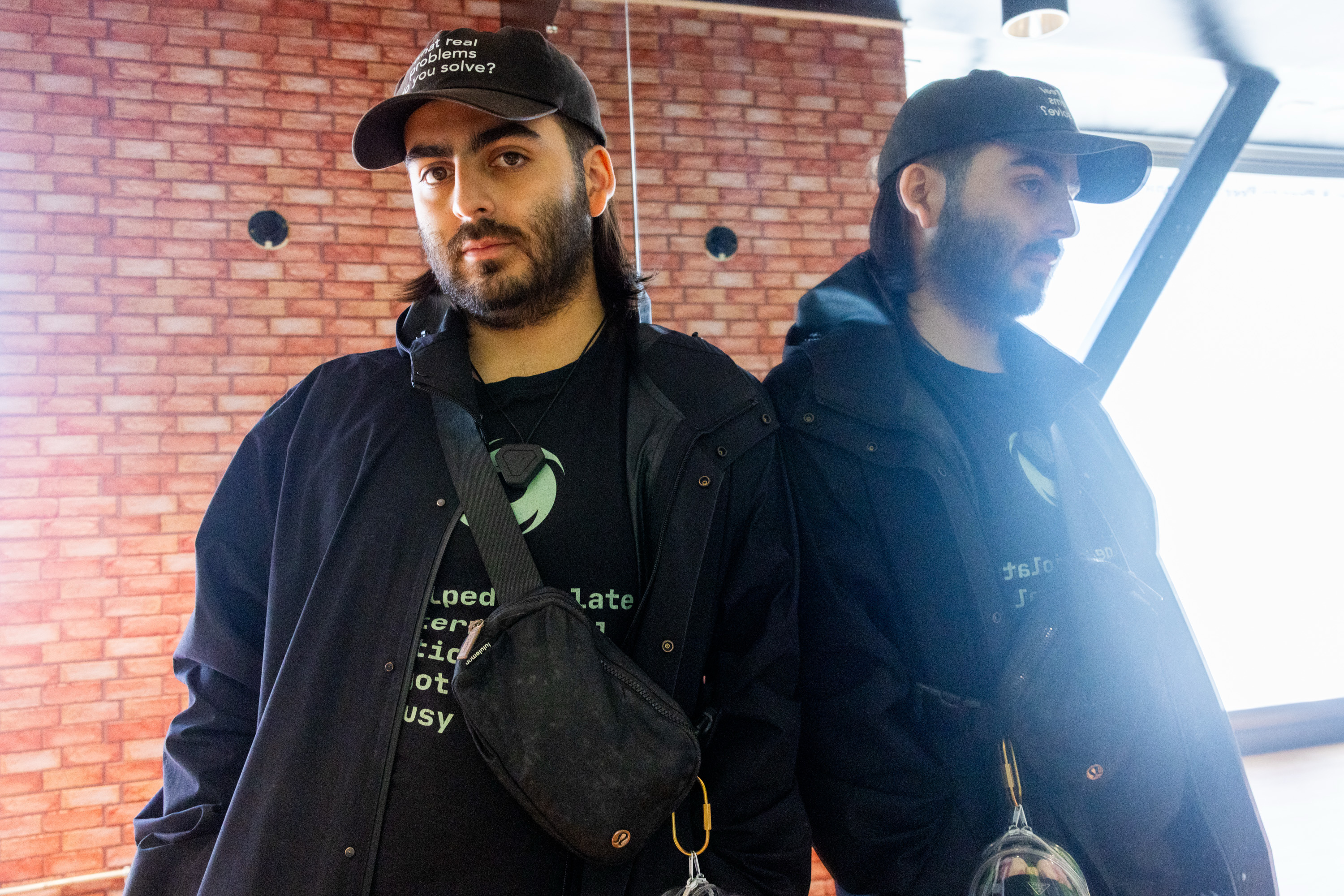 A man in dark clothing and a baseball cap stands in front of a brick wall. He's reflected in a mirror, wearing a crossbody bag and a black jacket.
