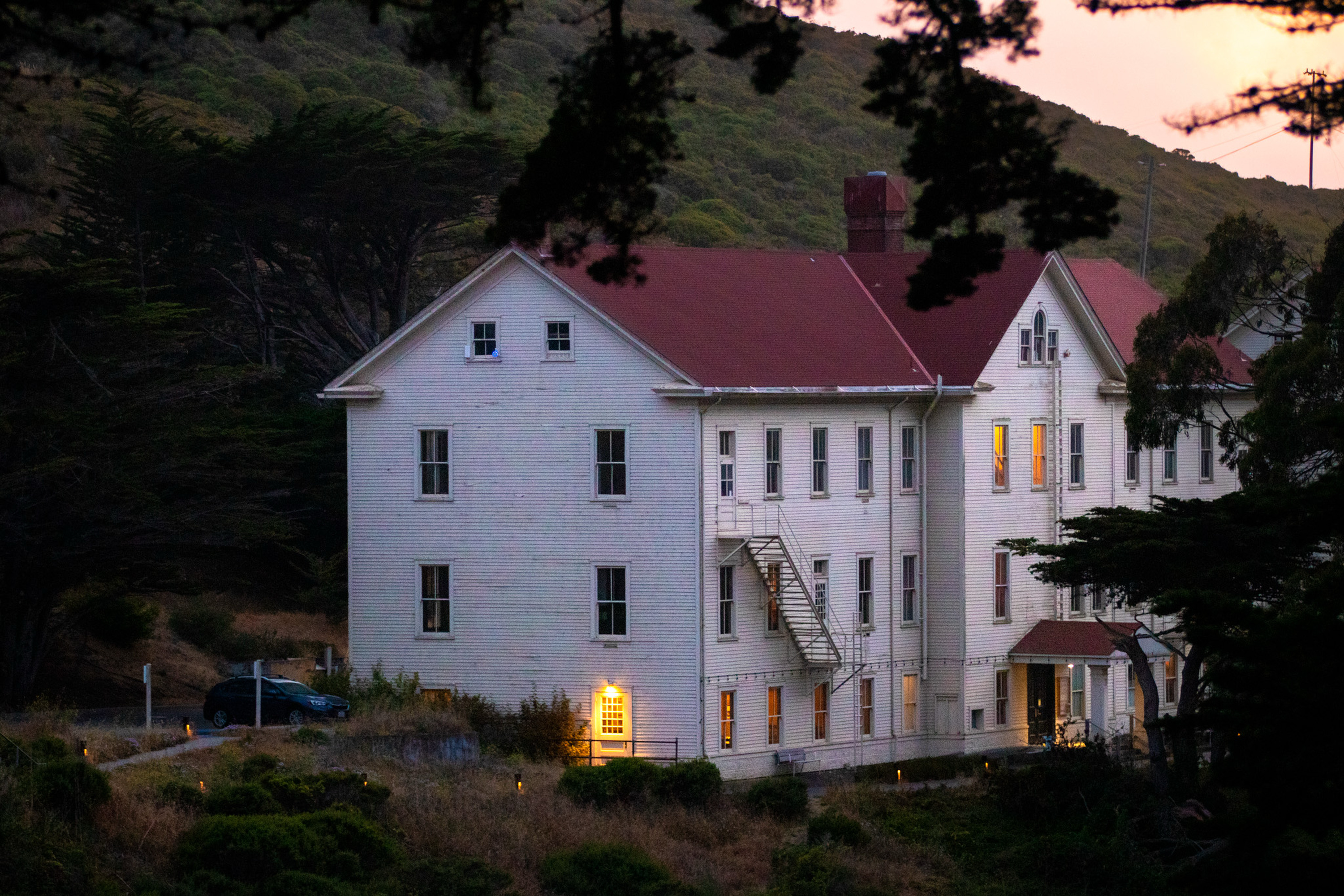 A large white, three-story building with a red roof is surrounded by trees. Some windows are warmly lit. An external staircase is visible, suggesting an emergency exit.