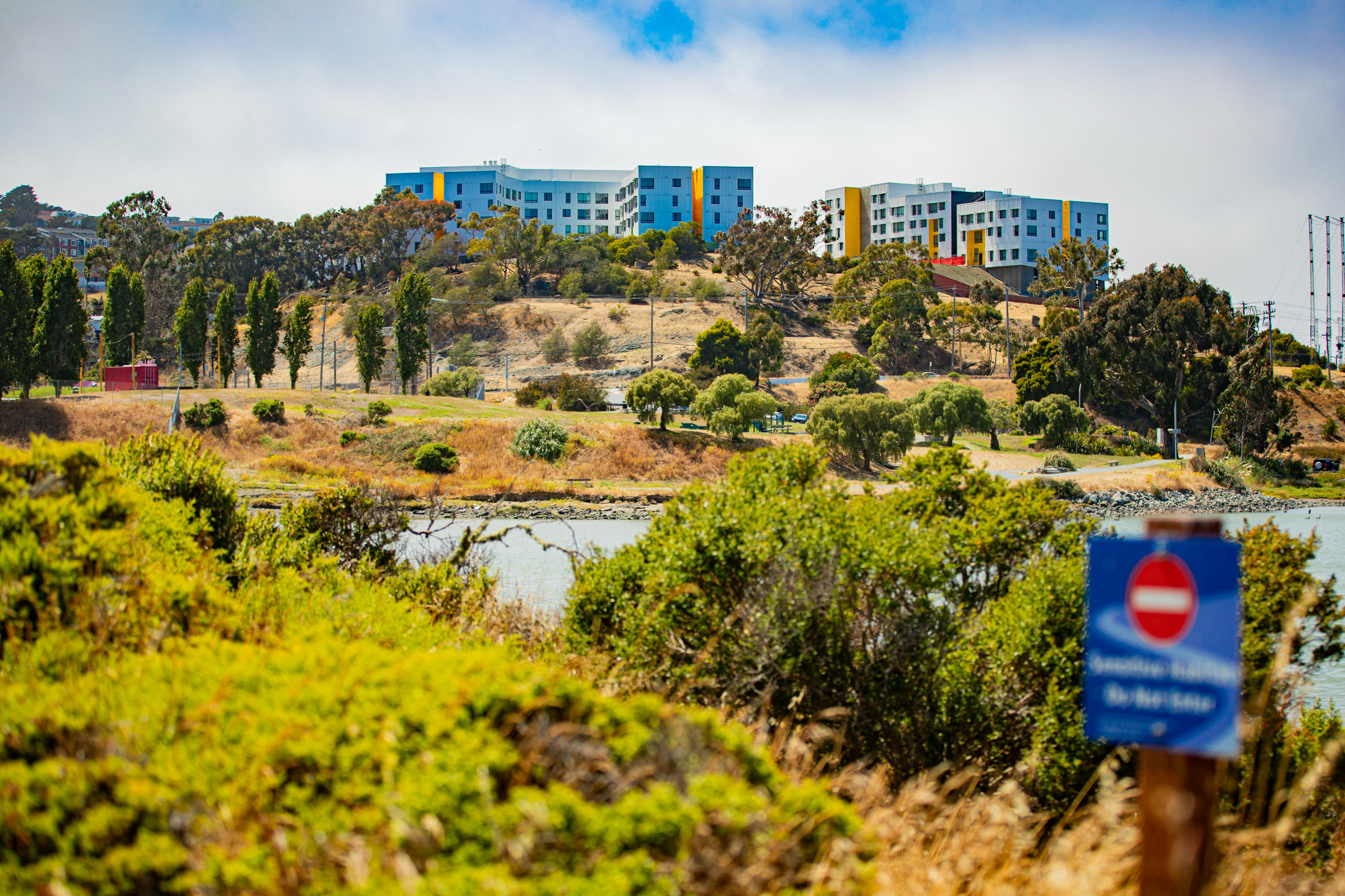 A hillside with colorful buildings at the top, surrounded by trees and vegetation. A body of water and a "Do Not Enter" sign appear in the foreground.