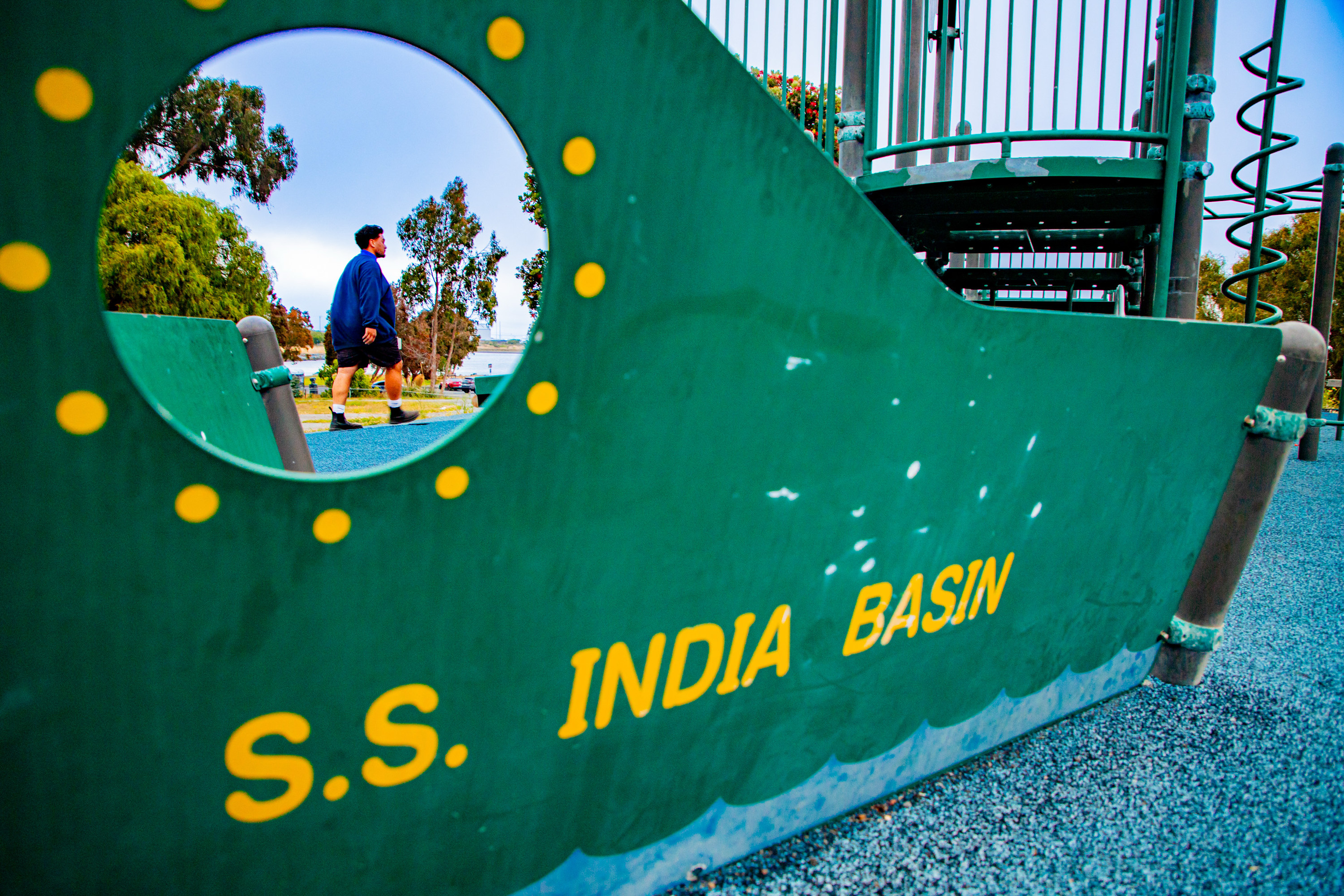 The image shows a green playground structure resembling a ship with &quot;S.S. INDIA BASIN&quot; written on it. A person walks past, seen through a circular cutout.
