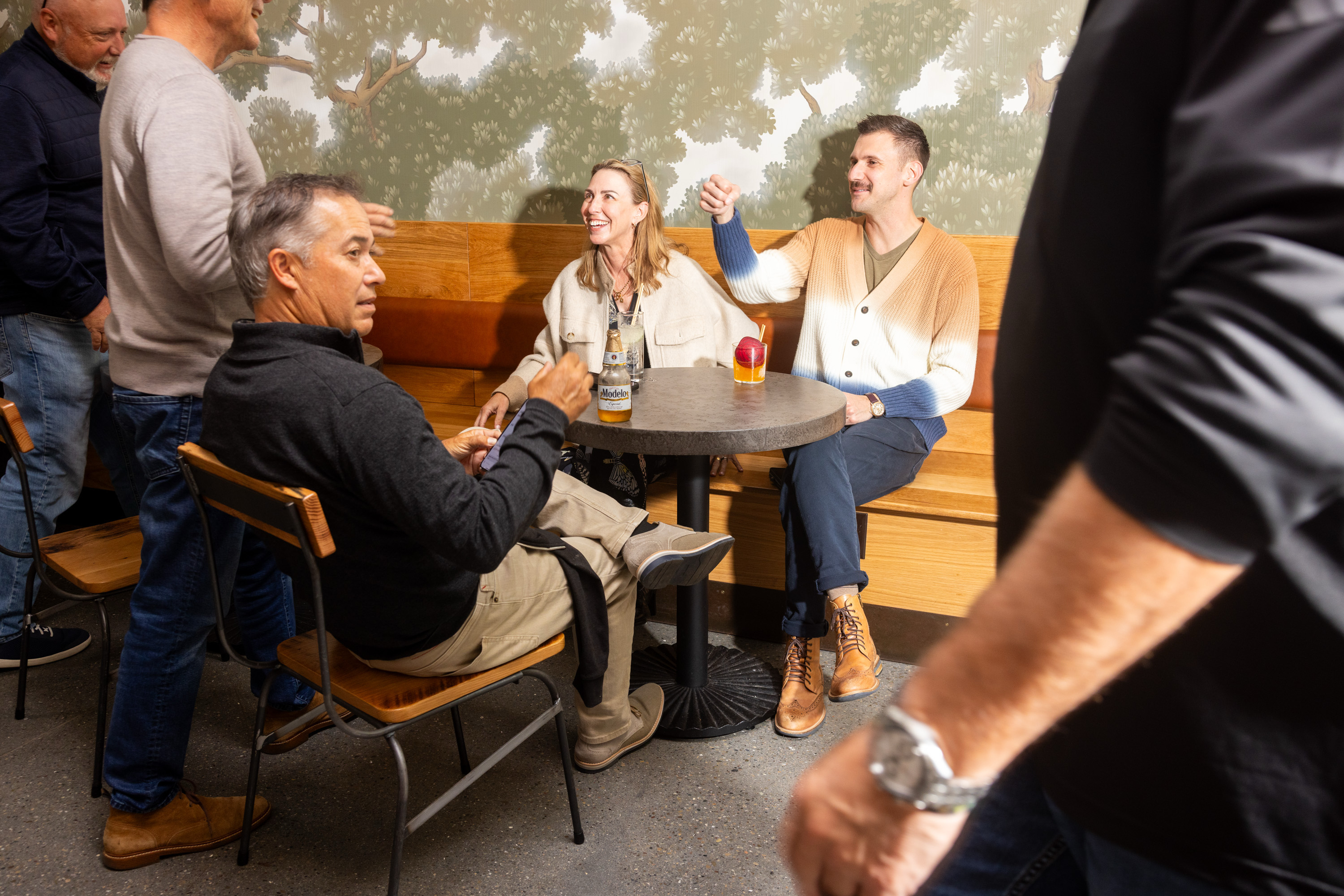 Several people are gathered in a casual indoor setting. Three are seated at a round table, with drinks, while others stand nearby, all engaged in conversation.