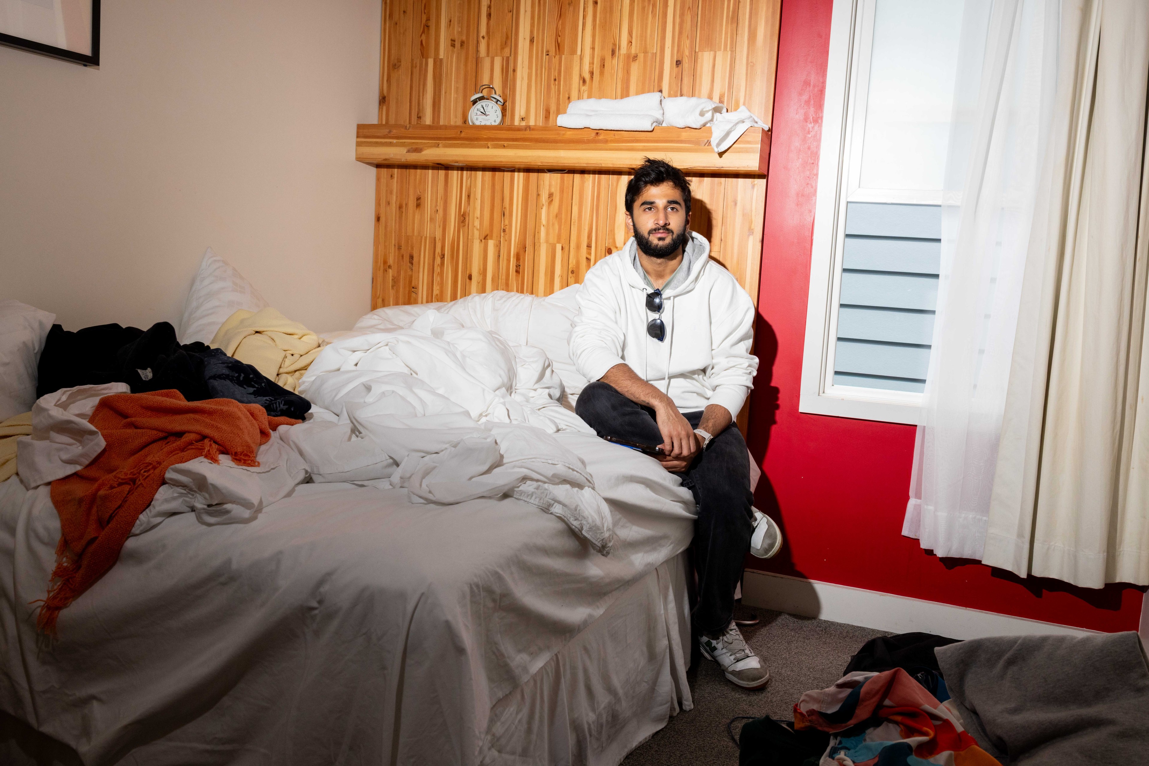 A person in a white hoodie sits on a bed with messy white sheets in a room. An alarm clock is on a shelf; clothes are scattered around the room.