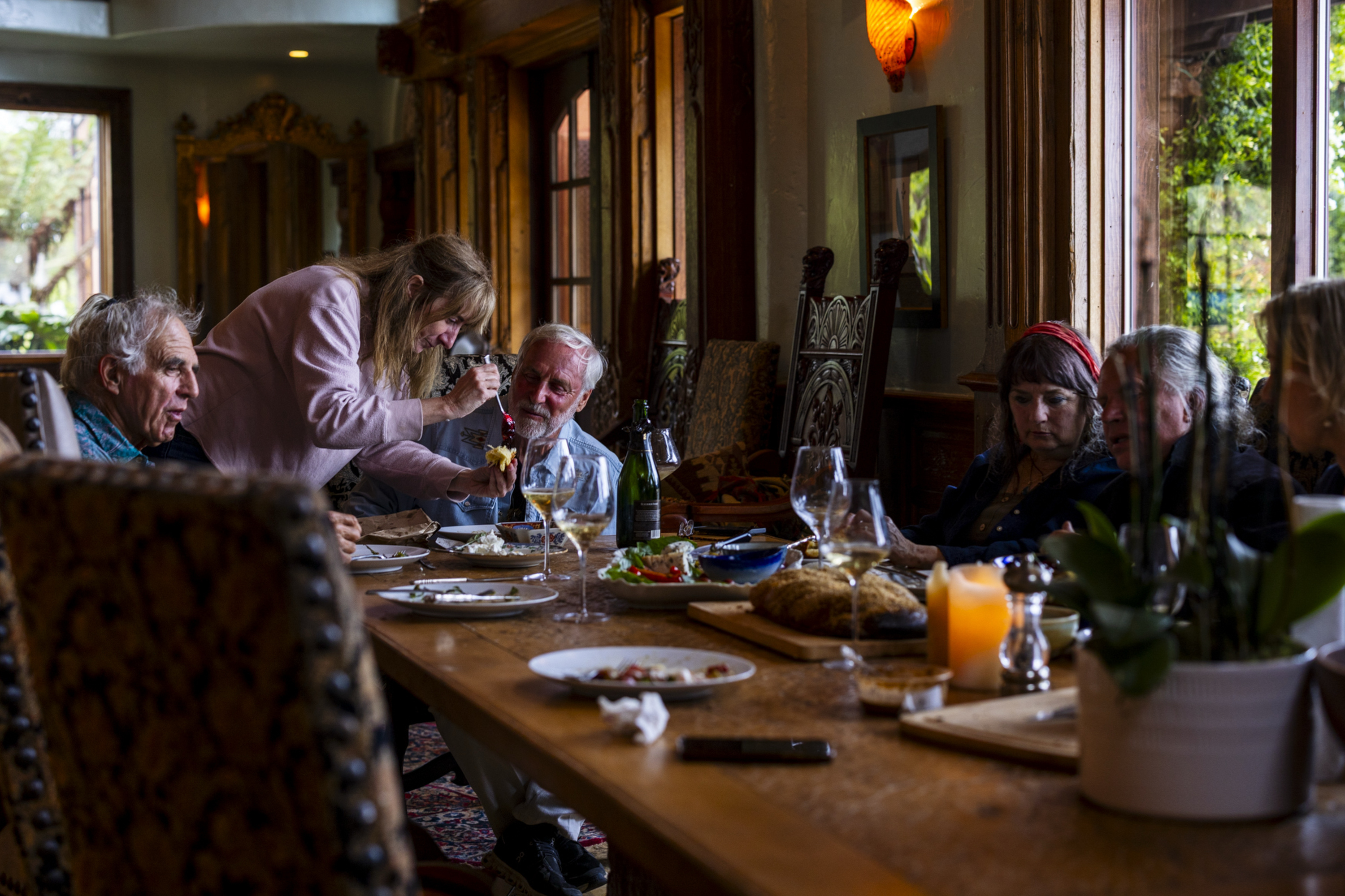 A group of six people is gathered around a dining table indoors. One woman is serving food to an elderly man. The table is set with dishes and a candle.