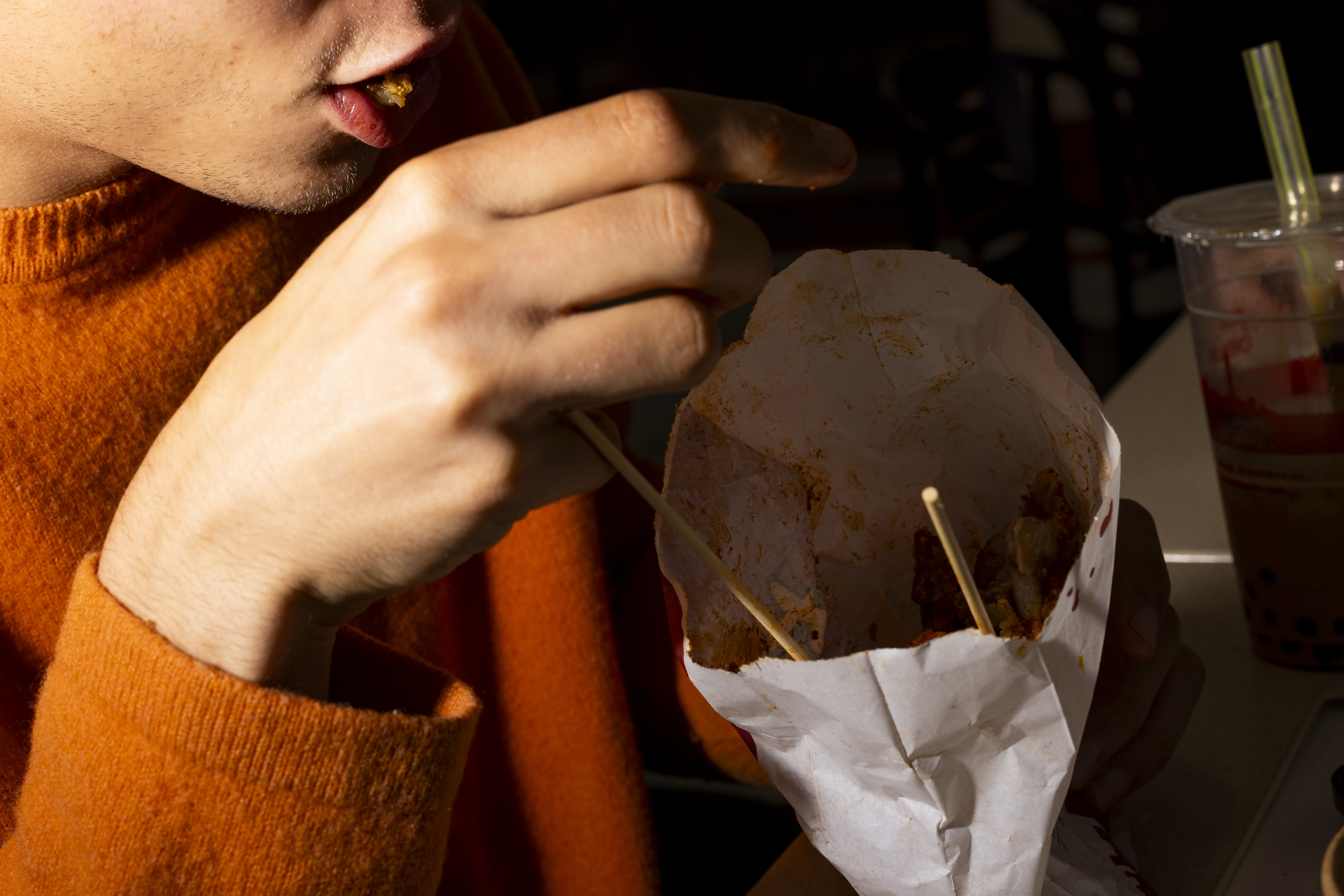 A person in an orange sweater eats food with chopsticks from a white paper bag, with bubble tea in a plastic cup next to them.