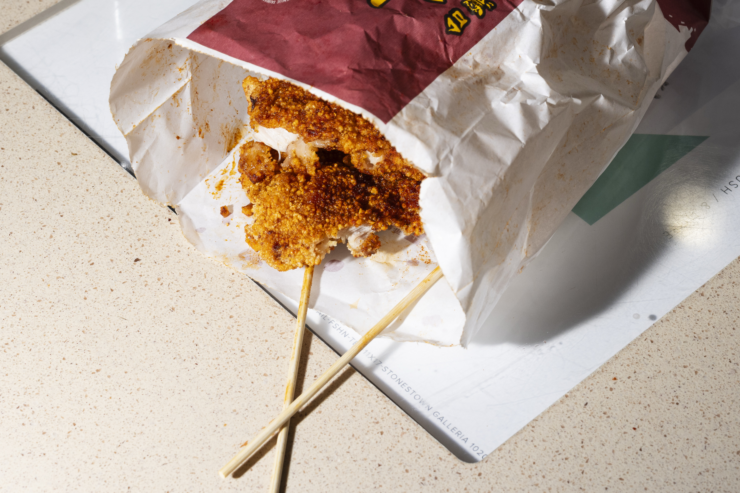 A paper bag lies on a table, partly open to reveal two skewers with crispy, spiced golden-brown fried chicken pieces inside.
