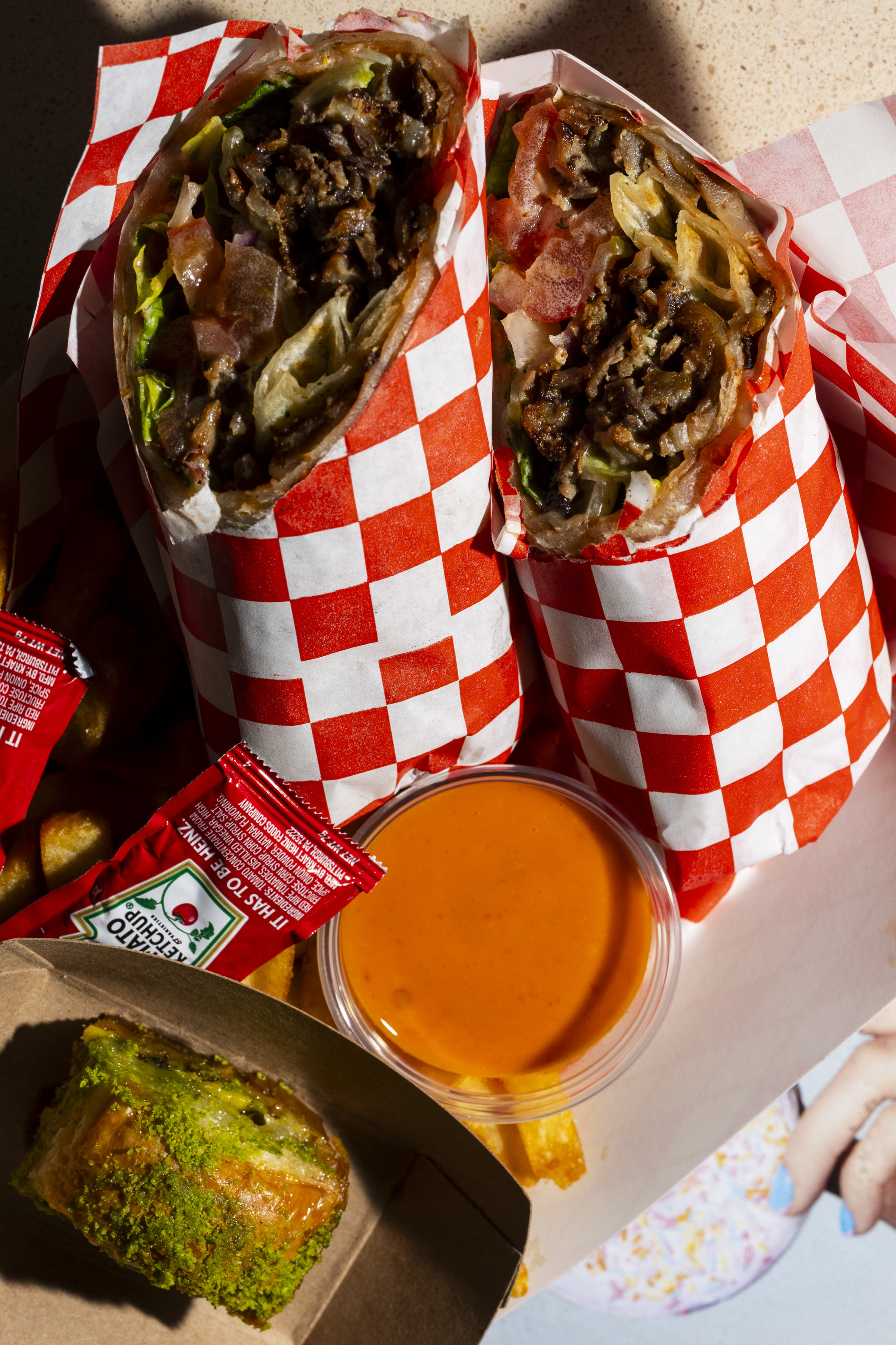 Two grilled wraps with meat and veggies are in red-checkered paper, accompanied by fries, ketchup packets, a container of orange sauce, and a pistachio dessert.