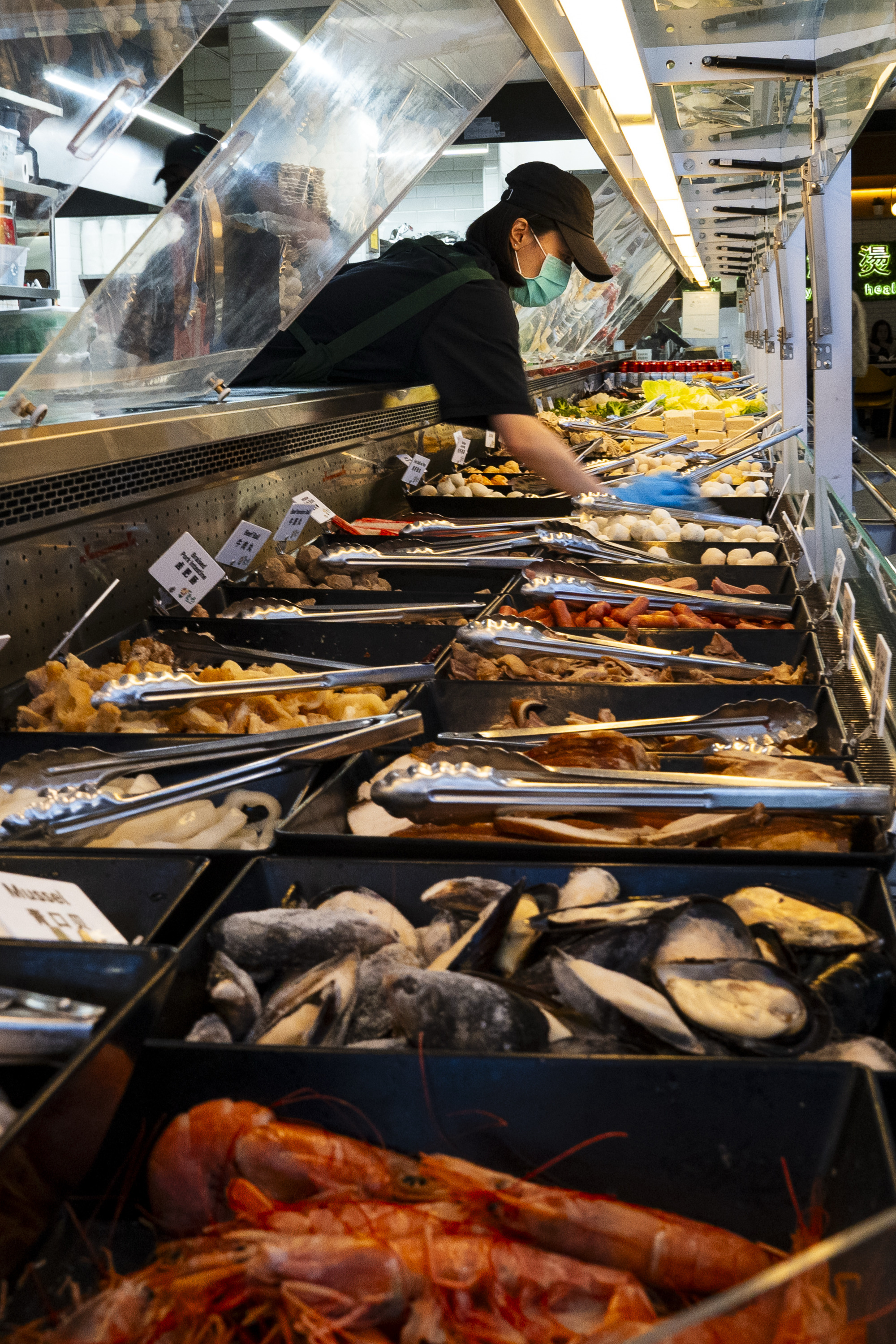 A masked worker arranges a variety of seafood and other fresh foods displayed in trays with tongs under a glass-covered market counter.