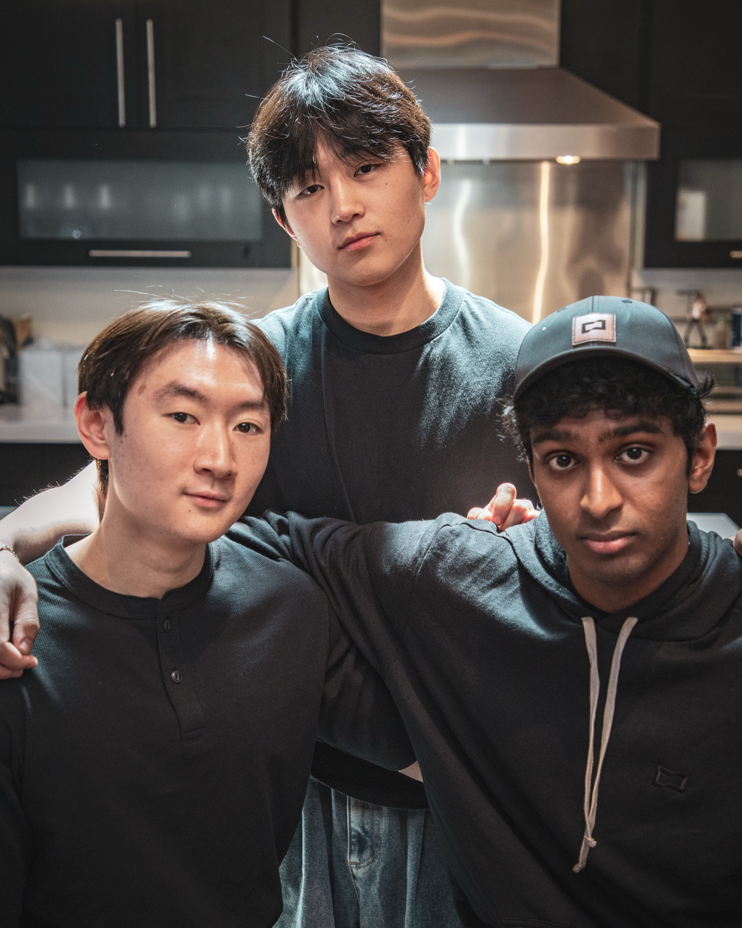 Three young men, wearing dark shirts, pose together in a kitchen. One stands while the other two sit, with arms around each other, under soft lighting.