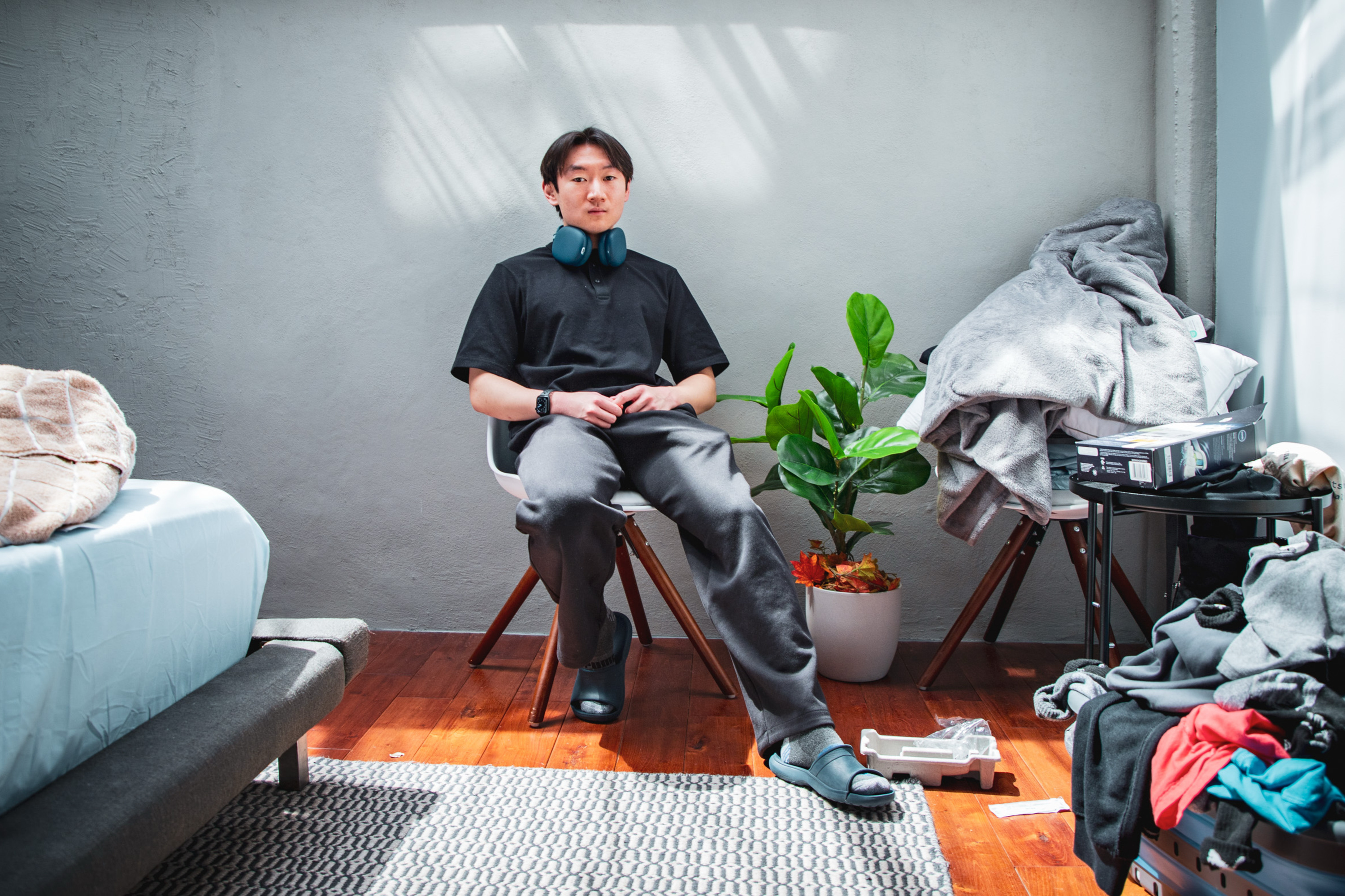 A person sits on a chair in a cluttered room with wooden floors. They're wearing a black shirt, gray pants, and headphones around their neck. Nearby, there's a plant and a messy pile of clothes.