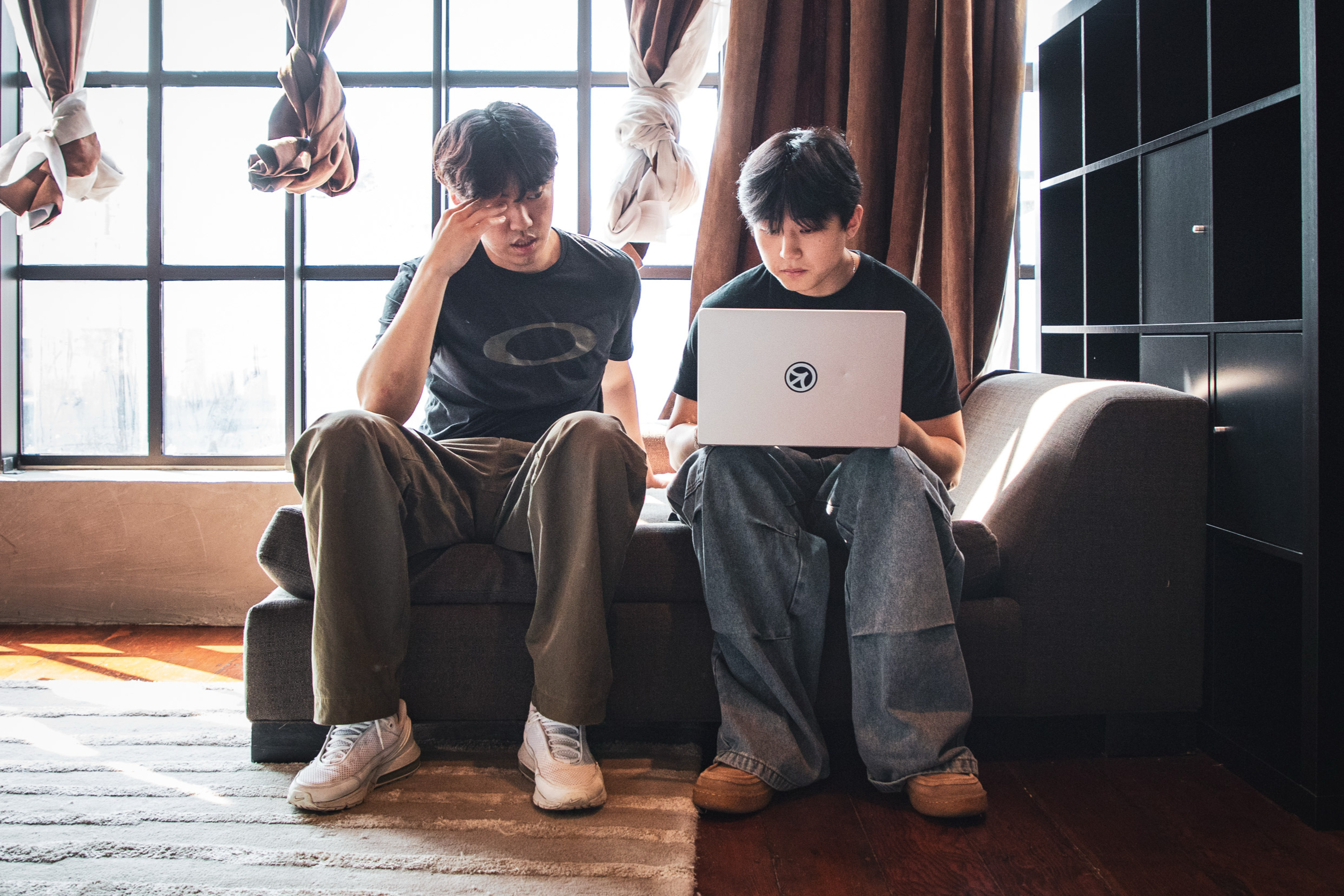 Two people are sitting on a couch by a large window. One appears thoughtful, while the other is focused on a laptop. The room is well-lit and cozy.