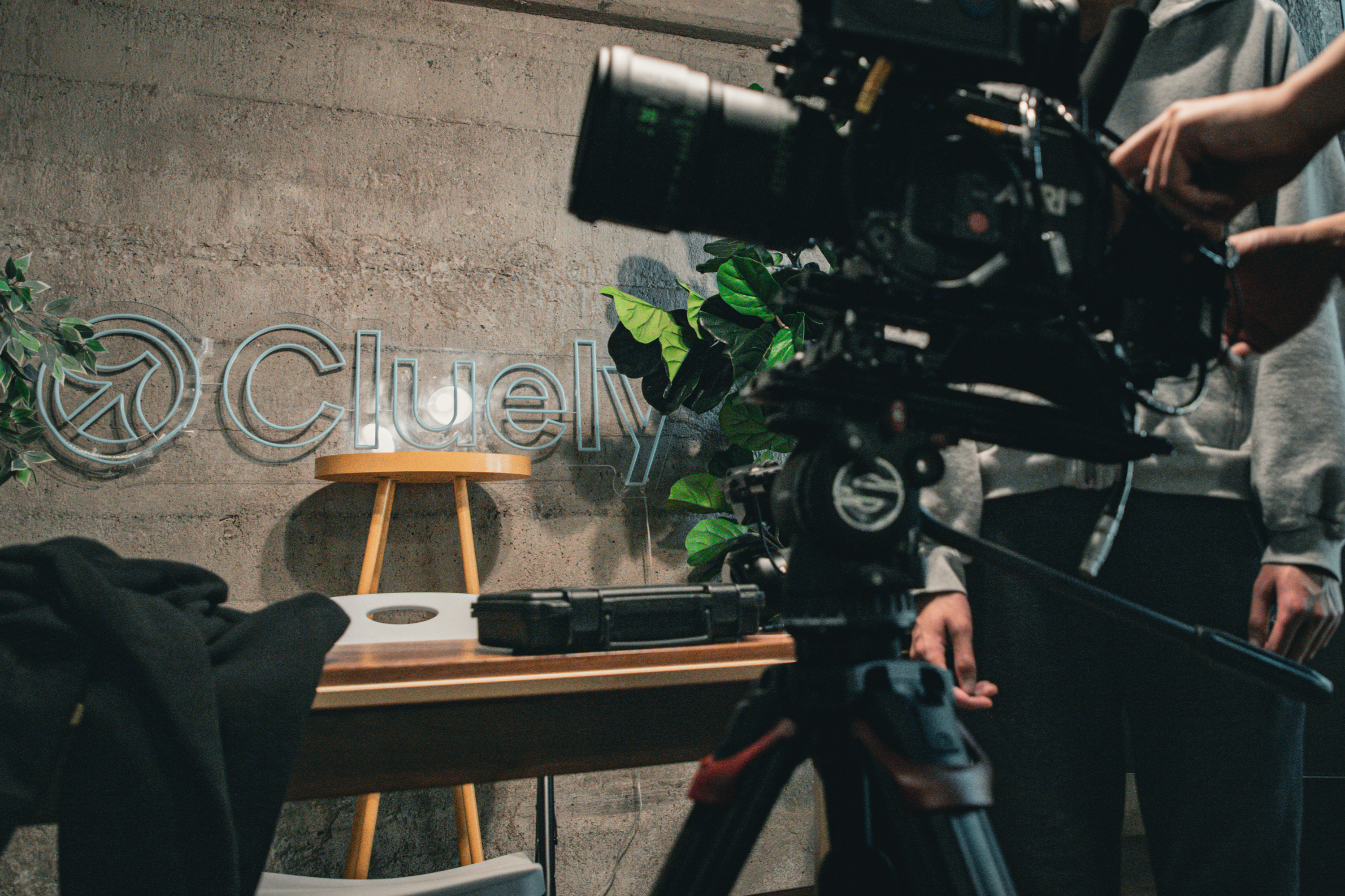 A professional camera setup is arranged in front of a table with a suitcase, chair, and a wall logo reading &quot;Cluely&quot; against a concrete background with plants.