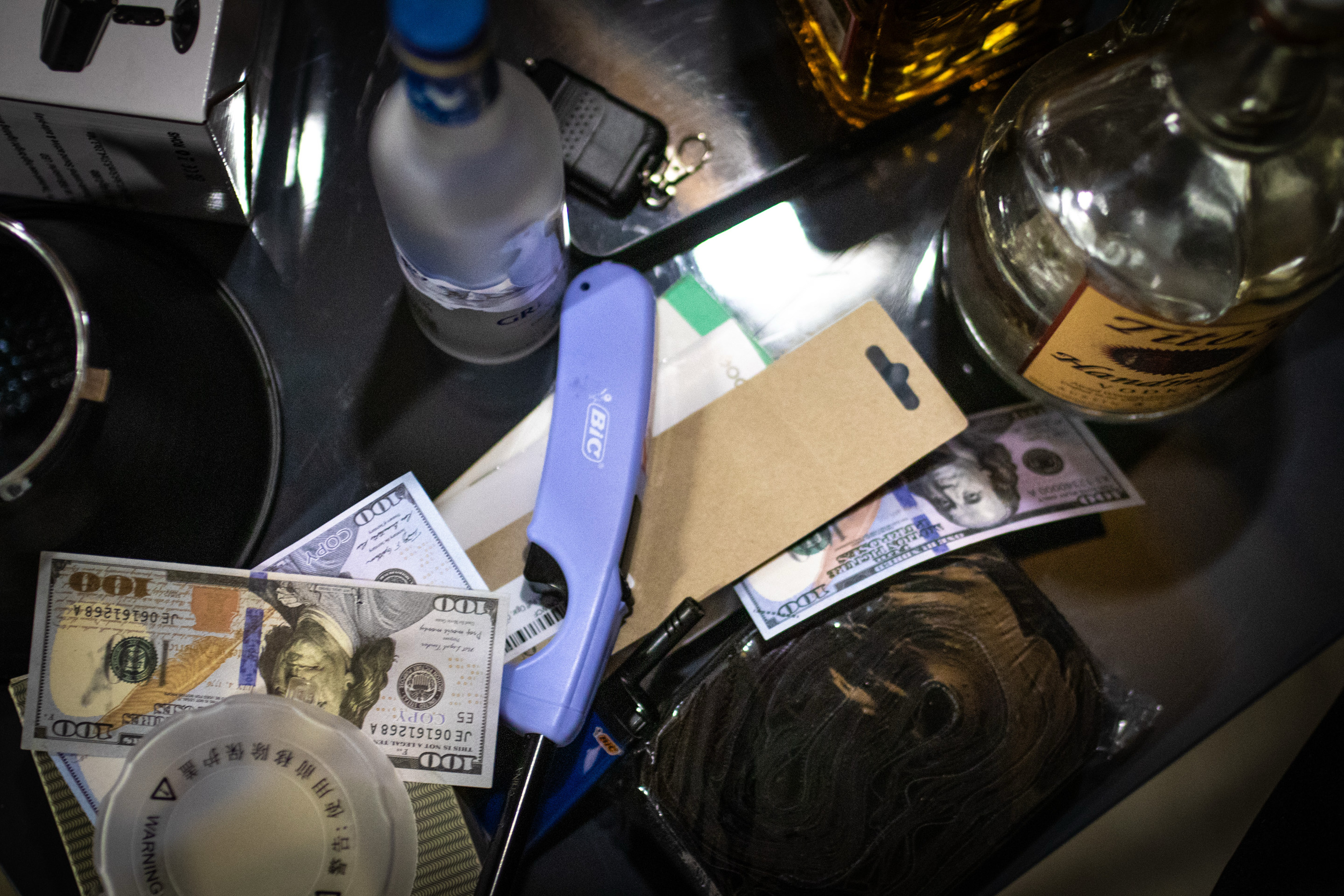 The image shows bottles, fake money, a lighter, keys, and some small items on a table, suggesting a casual setting with various objects scattered around.