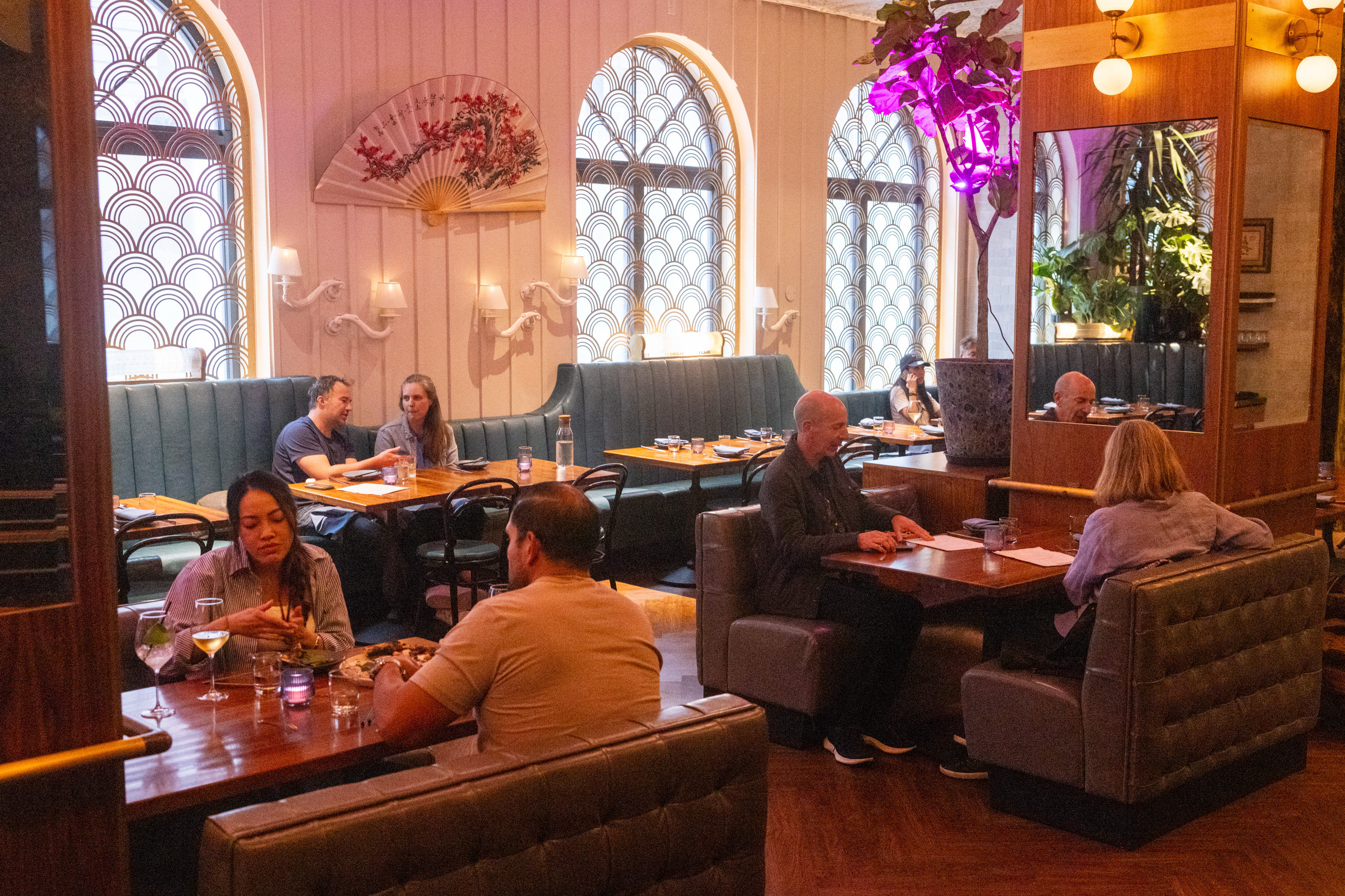The image shows a cozy restaurant with people dining in semi-circular booths. The decor includes arched windows, a large fan on the wall, and warm lighting.