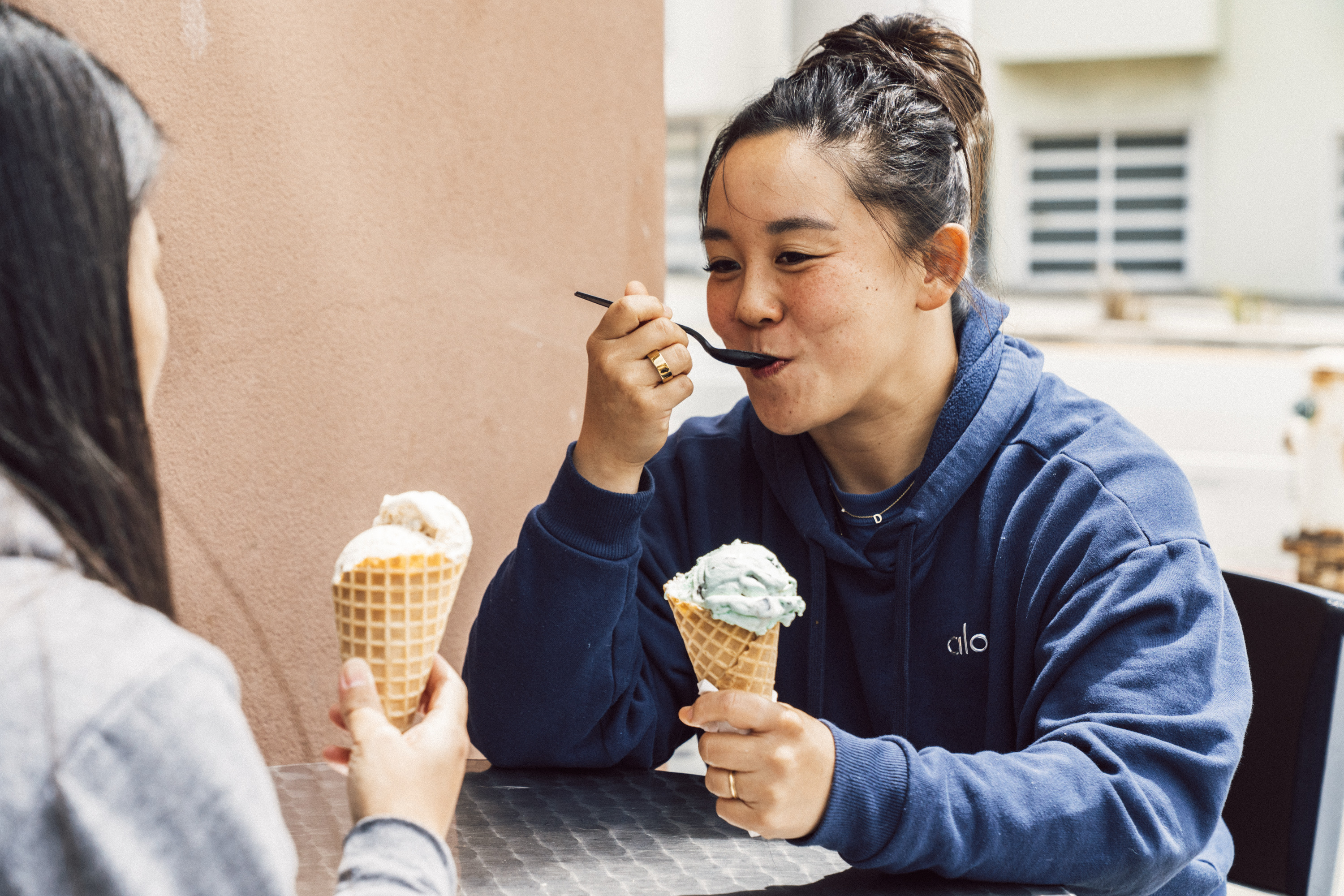 Two people are sitting at an outdoor table enjoying ice cream cones. One is using a spoon, wearing a blue hoodie, while facing a friend in a gray top.