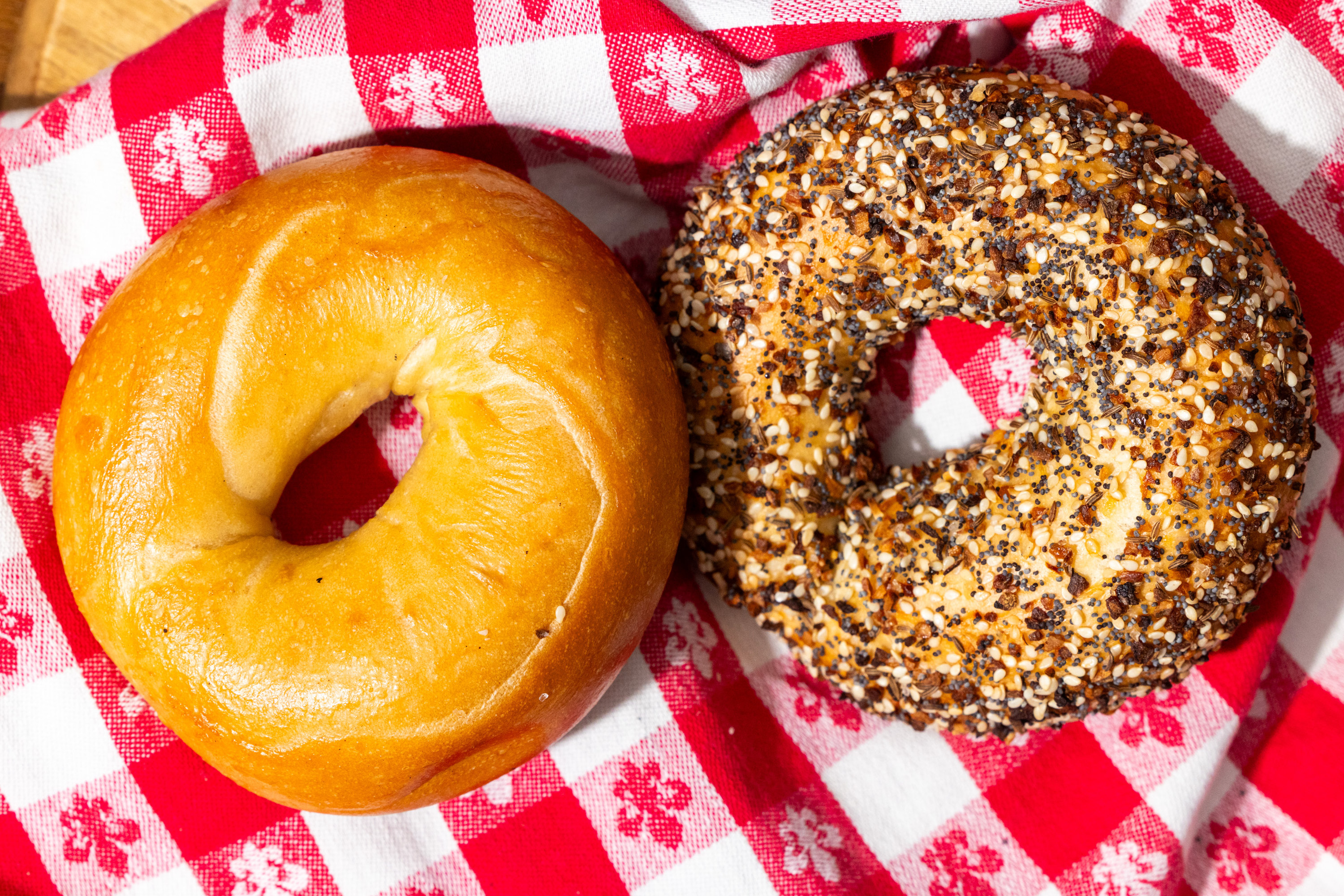 Two bagels sit on a red and white checkered cloth: one plain, golden-brown, and the other covered in a variety of seeds and seasonings.