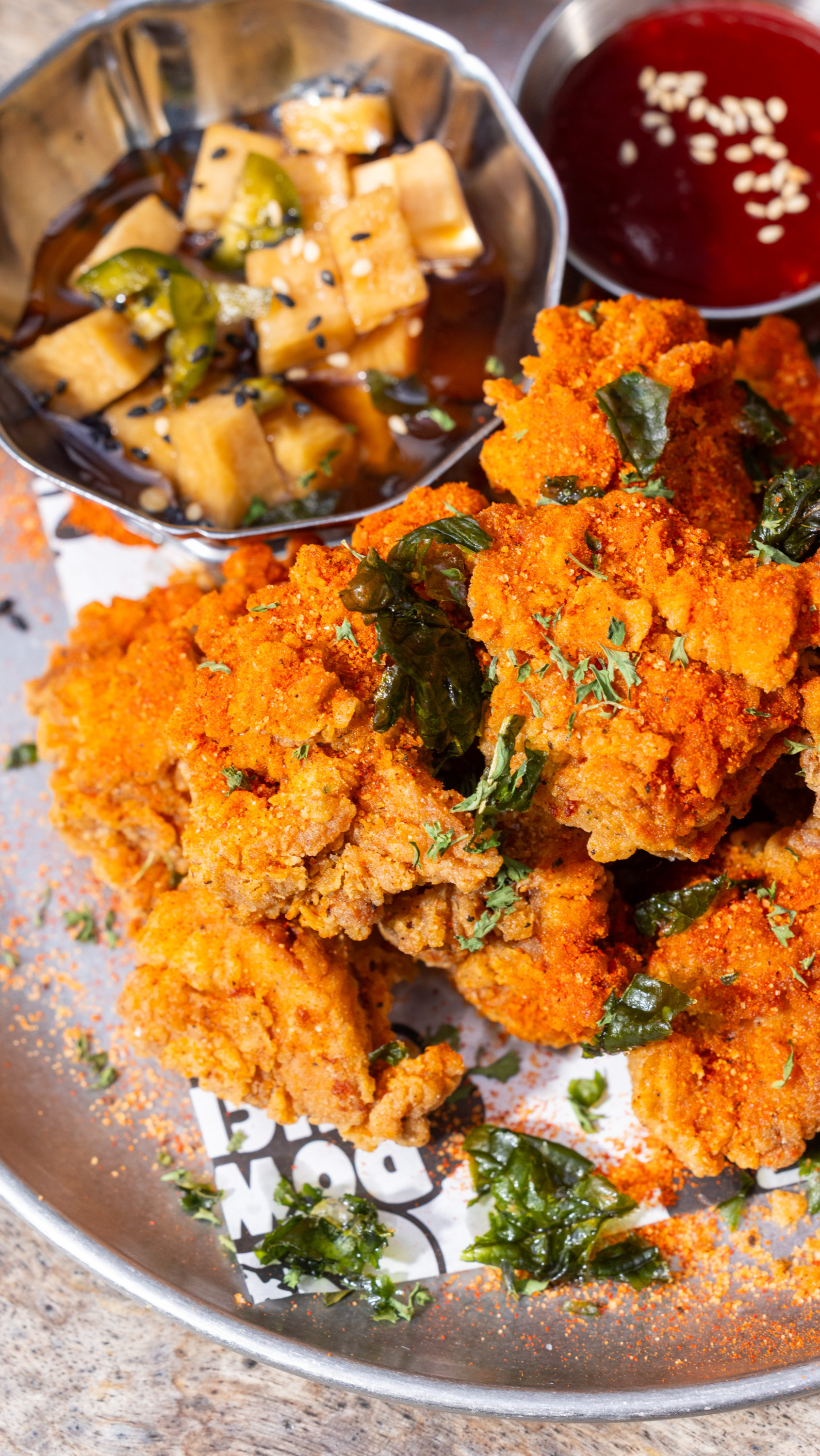 Crispy fried chicken with orange seasoning is garnished with herbs. It's served with small bowls of bright red sauce with sesame seeds and pickled cubes.