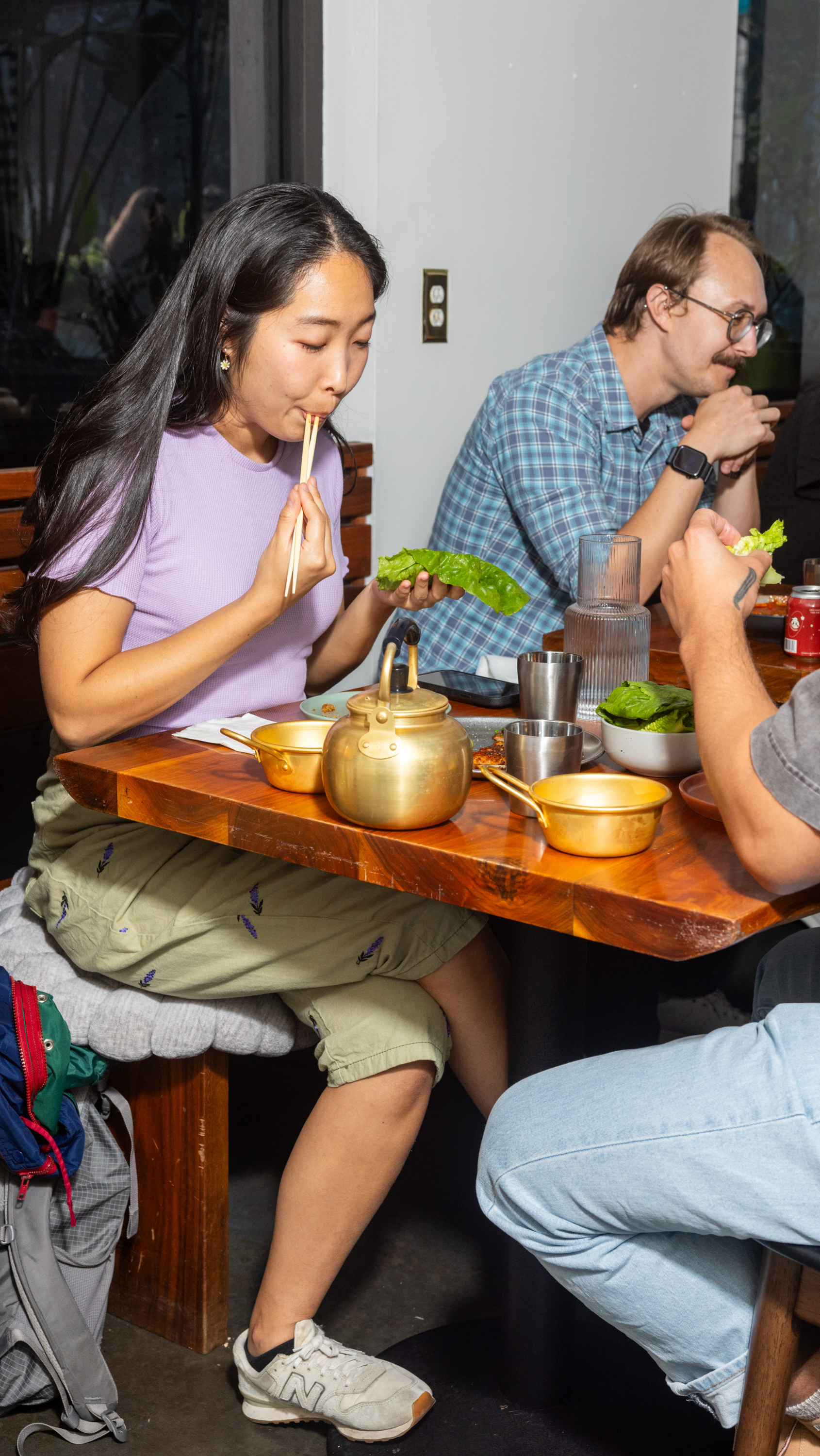 A group of people is dining at a wooden table. One person is eating with chopsticks, holding a leaf, while others converse. A teapot and bowls are on the table.