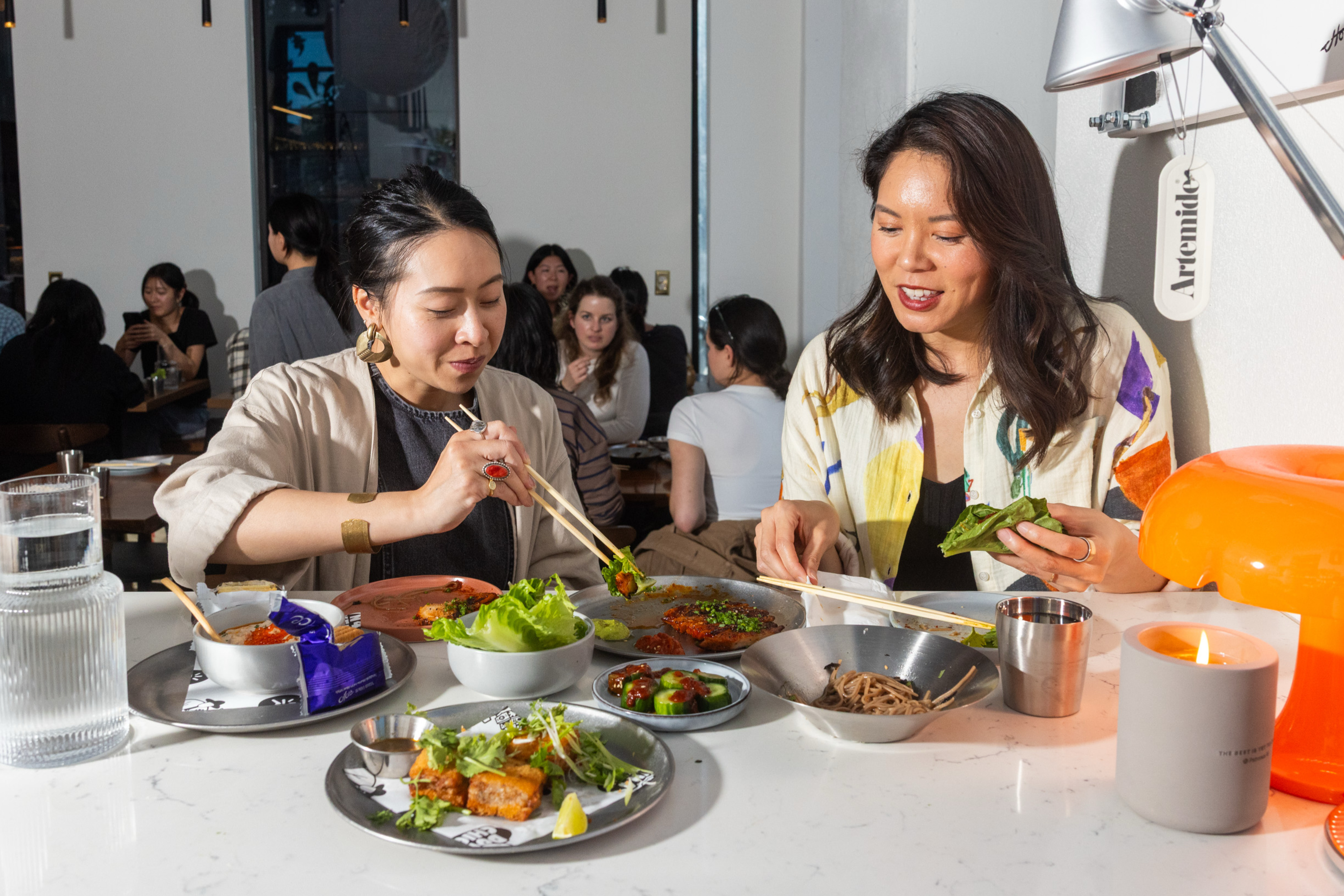 Two women sit at a table enjoying a meal with various dishes. They're using chopsticks, surrounded by plates of food, a candle, and a bright orange lamp.