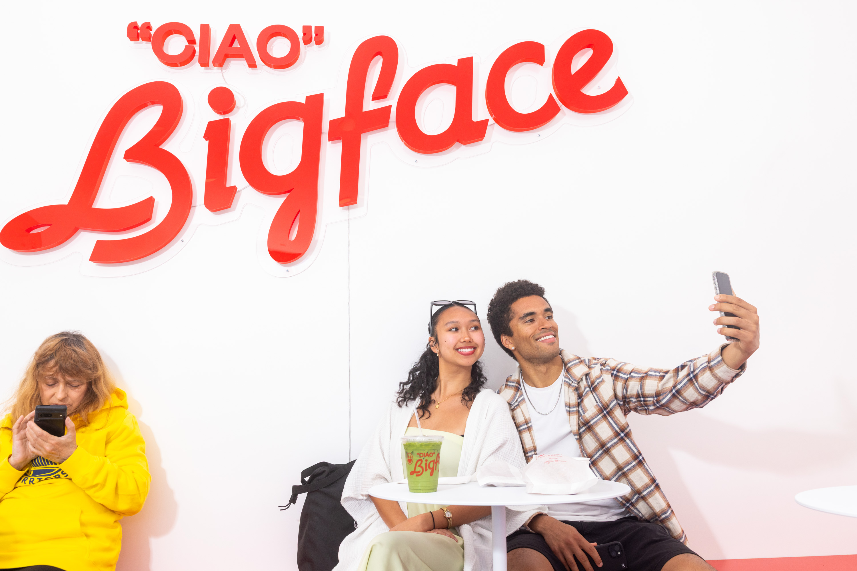 A couple takes a selfie under a red "Ciao Bigface" sign. They are sitting at a small table, and a person in a yellow hoodie is on their phone to the left.